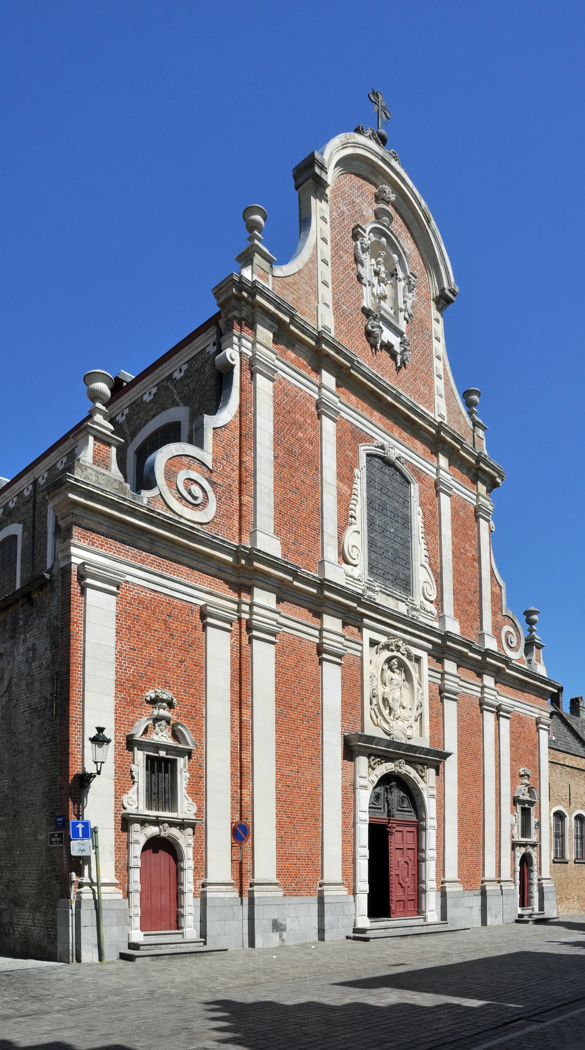 Fachada de iglesia barroca de ladrillo con pilastras de piedra blanca, puertas rojas, cartuchos decorativos y una cruz sobre el frontón contra un cielo azul despejado.