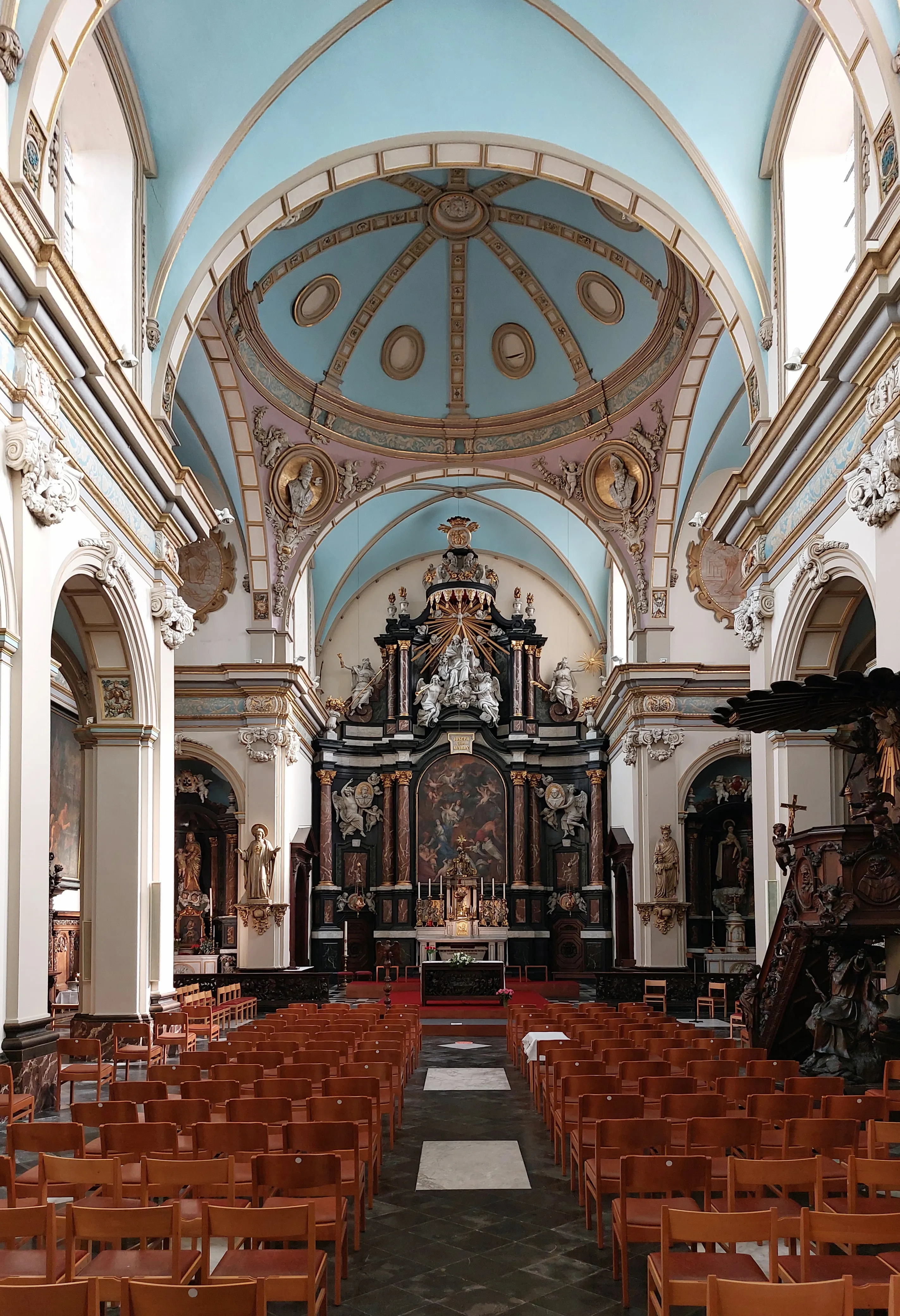 Interior de una iglesia barroca con techo abovedado azul, altar ornamentado en negro y oro, y filas de sillas de madera frente al santuario.