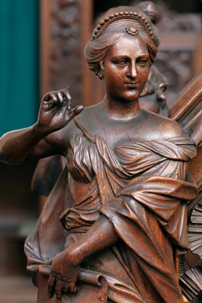 Primer plano de una escultura de madera tallada de una figura femenina coronada con drapeados clásicos fluidos, que sostiene una pluma de ave con libros tallados cerca