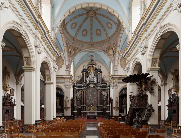Interior de la nave de una iglesia con hileras de bancos de madera, columnas blancas ornamentadas con arcos, un techo abovedado azul con medallones decorativos y un elaborado altar oscuro al fondo.