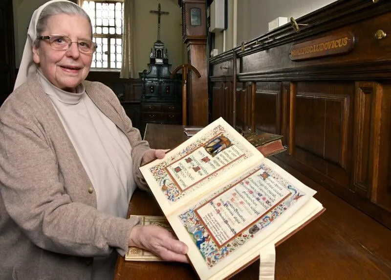 Una monja carmelita con hábito beige y gafas sostiene abierto un manuscrito iluminado con páginas decoradas, de pie en una histórica habitación con paneles de madera y un crucifijo en la pared.