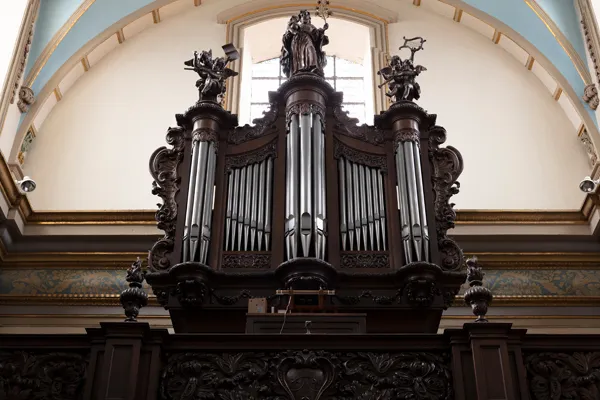 Ornato órgano de madera oscura con estatuas talladas en la parte superior y tubos de plata, situado contra un techo arqueado claro con detalles azules dentro de una iglesia.
