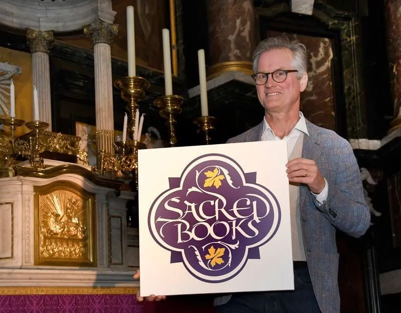 Un hombre con blazer sostiene un cartel blanco con el logotipo morado y dorado de Sacred Books frente a un altar de iglesia con velas altas y columnas de mármol.