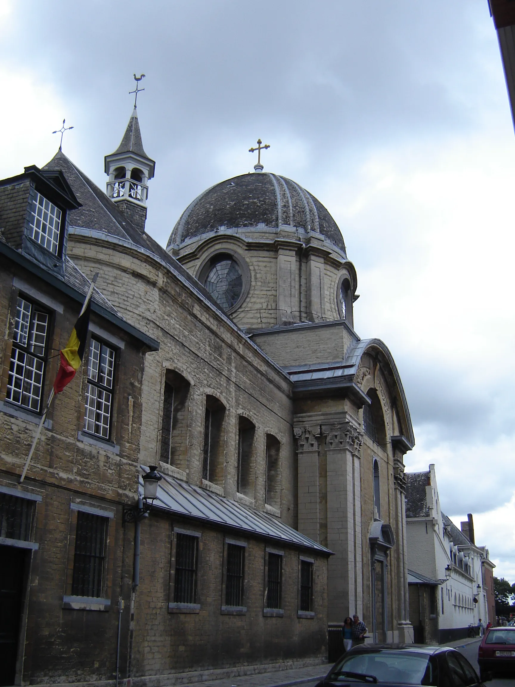 Vista exterior del Convento Inglés en Brujas que muestra una iglesia con cúpula y cruz, fachada de ladrillo con ventanas arqueadas, una pequeña torre de campana con una veleta y una bandera belga.