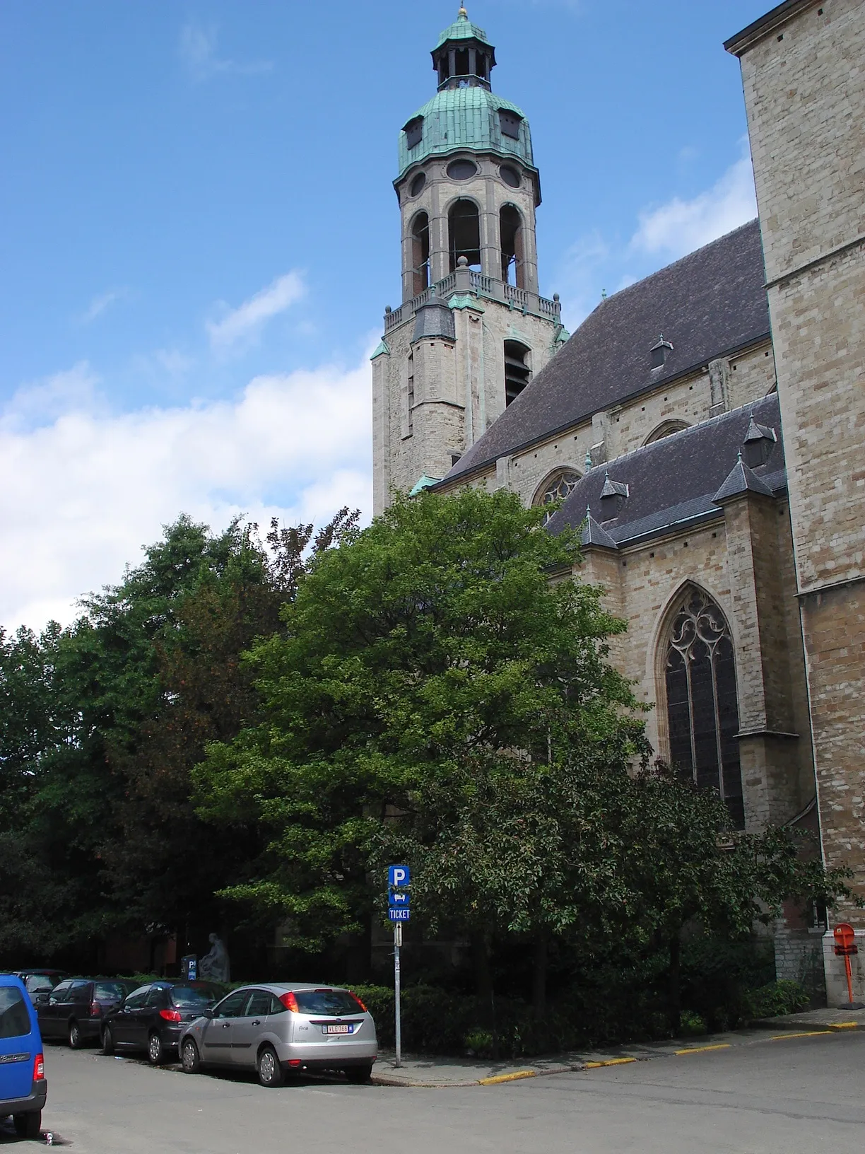 Vista exterior de la Saint Andrew's Church que muestra su alta torre de piedra con cúpula de cobre verde, ventanas góticas y árboles circundantes con coches aparcados en primer plano.