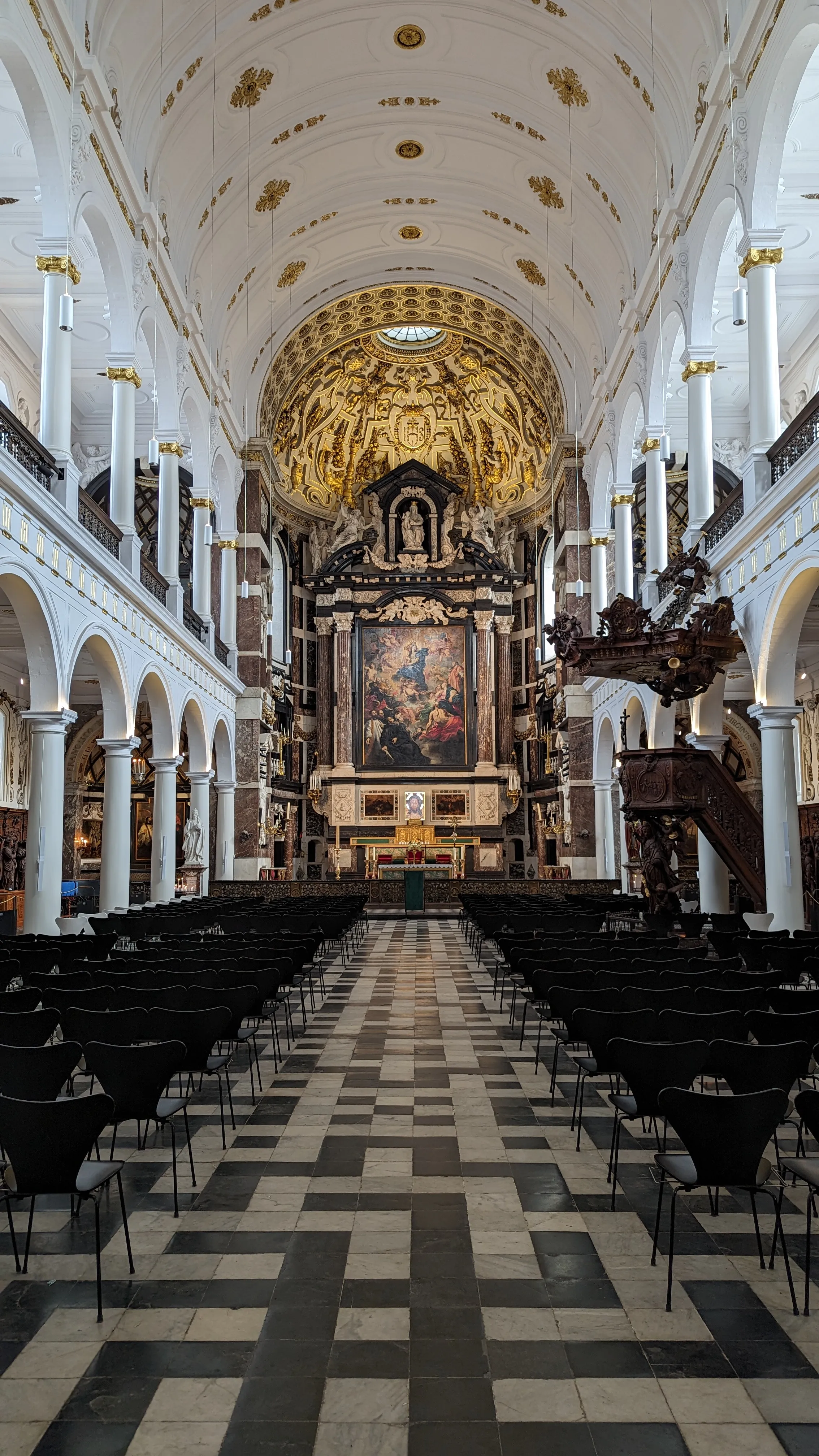 Interieur van de Sint-Carolus Borromeüskerk met een geblokte marmeren vloer, witte zuilen, een verguld plafond en een sfeervol altaar met een groot schilderij.