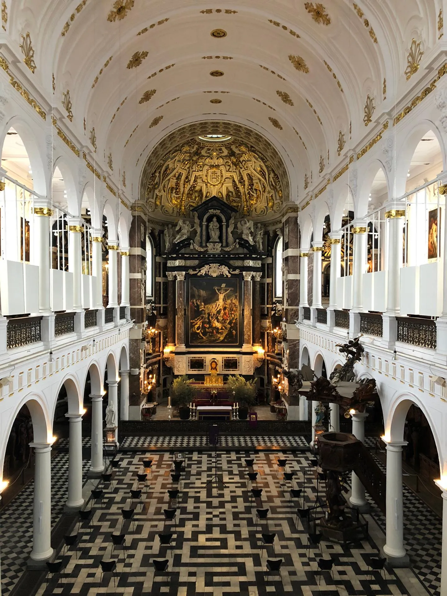 Interieur van een barokke kerkbeuk met witte boogzuilen, een goudversierd gewelfd plafond, een sierlijk altaar met een groot schilderij en een zwart-witte geometrische tegelvloer