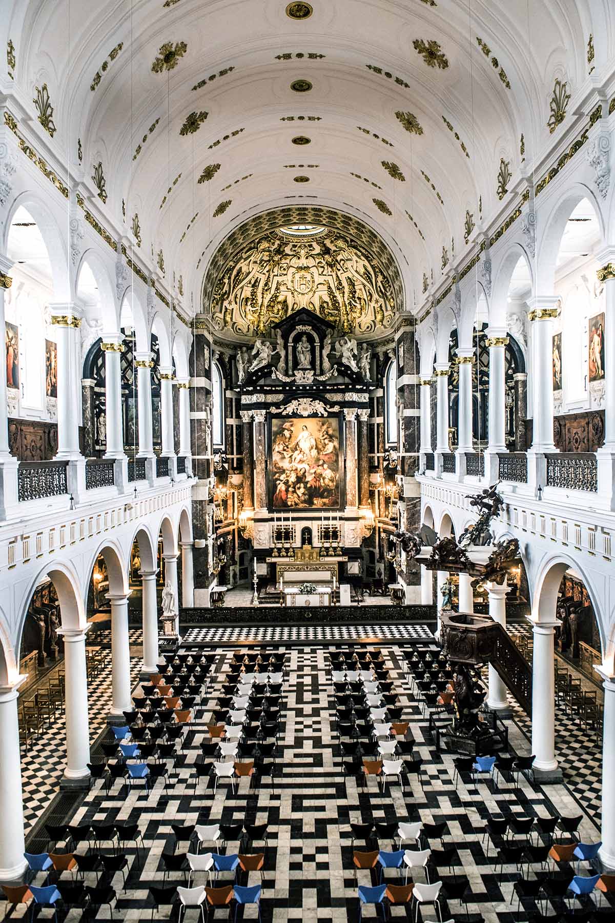 Interieur van het kerkschip met een zwart-witte geometrische marmeren vloer, witte zuilen en een rijkversierd altaar onder een gewelfd plafond.