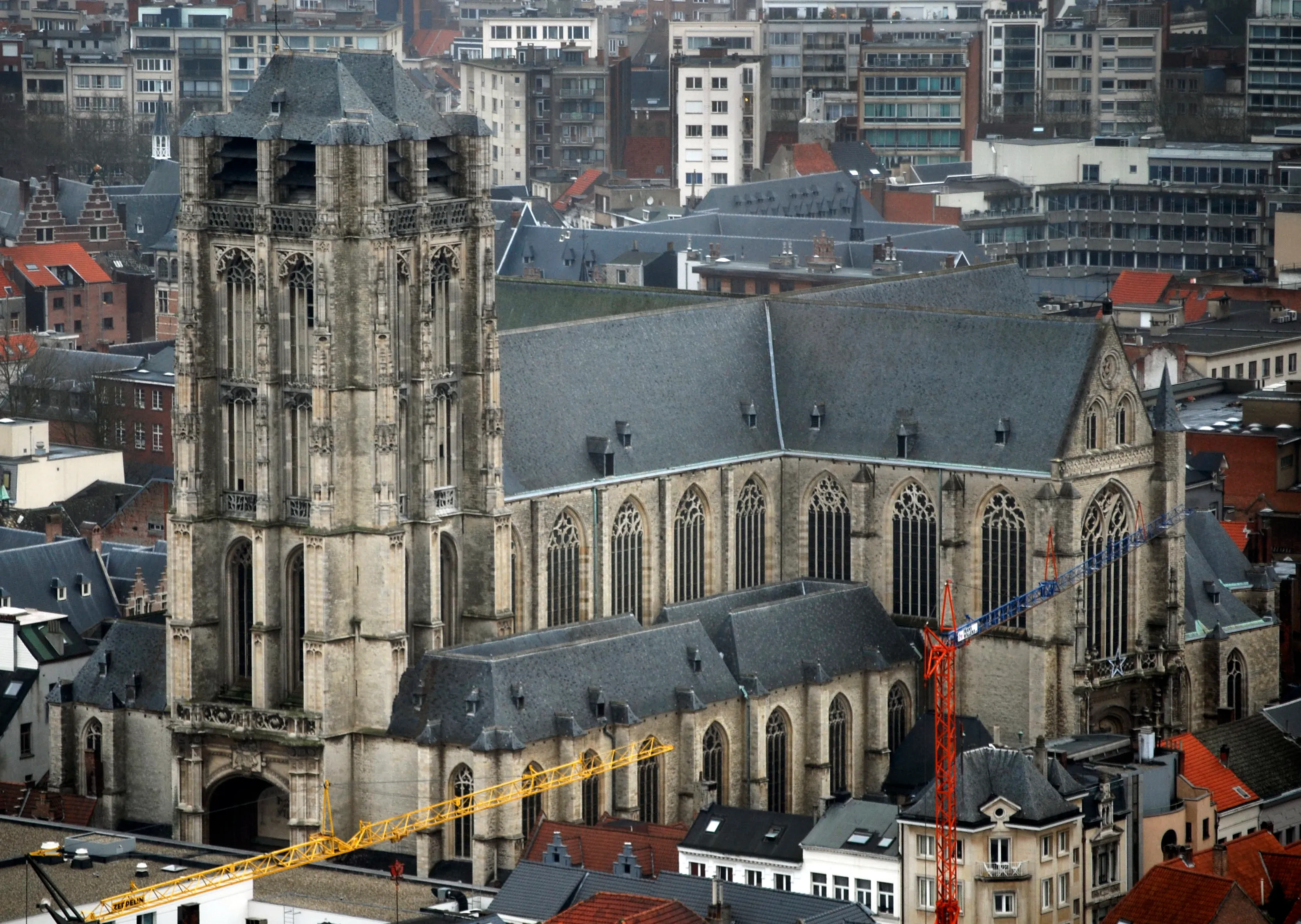 Vista aérea de la Iglesia de Santiago en Amberes que muestra su alta torre gótica, el tejado de pizarra, las ventanas góticas y el paisaje urbano circundante con grúas de construcción amarillas y naranjas.