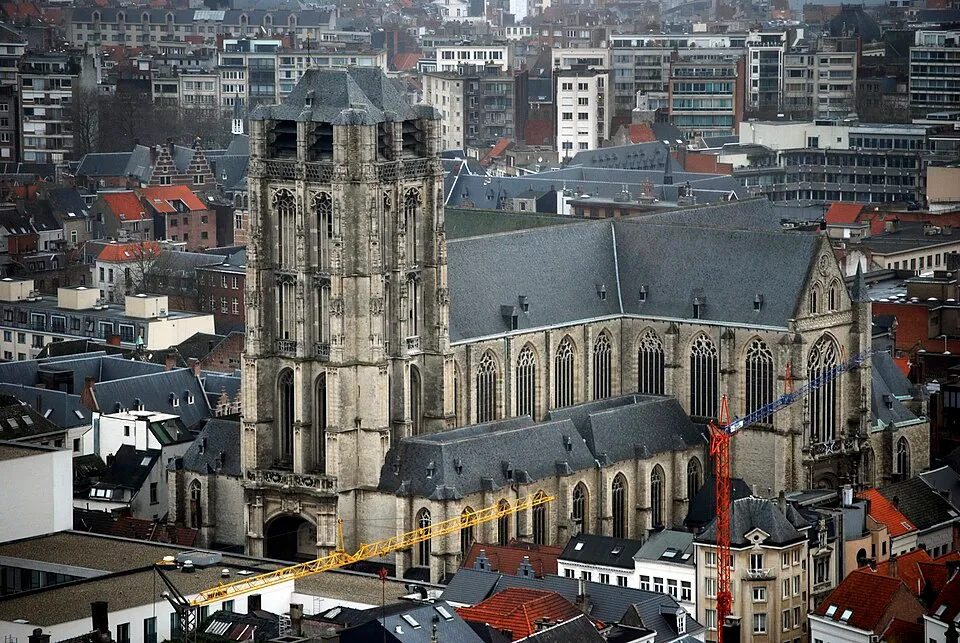 Vista aérea de la Iglesia de Santiago que muestra su alta torre gótica y su larga nave con ventanas de arco apuntado, rodeada por la ciudad de Amberes con grúas de construcción en primer plano