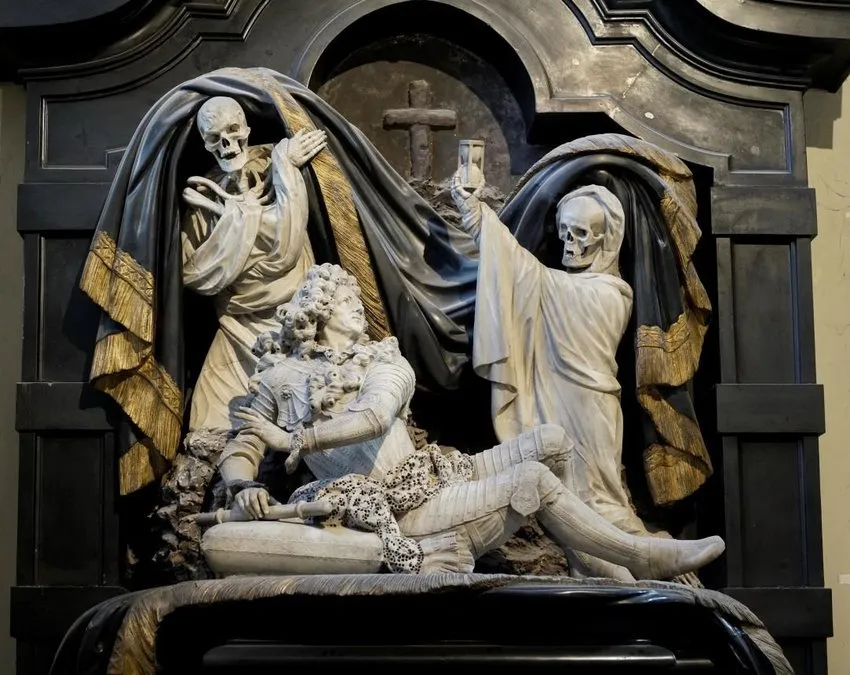 Escultura de monumento funerario de mármol que muestra dos figuras de esqueleto apartando un cortinaje oscuro con flecos dorados para revelar una figura yacente con armadura, con una cruz visible en la hornacina del fondo.