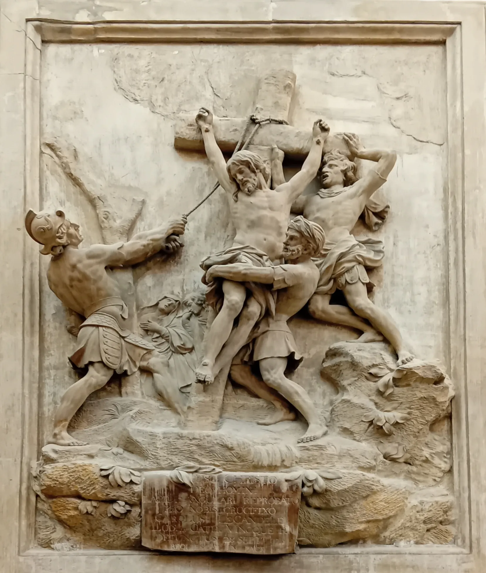 Relieve de piedra que muestra varias figuras elevando a Jesús a una cruz, con un soldado romano a la izquierda y una placa con una inscripción en la base.