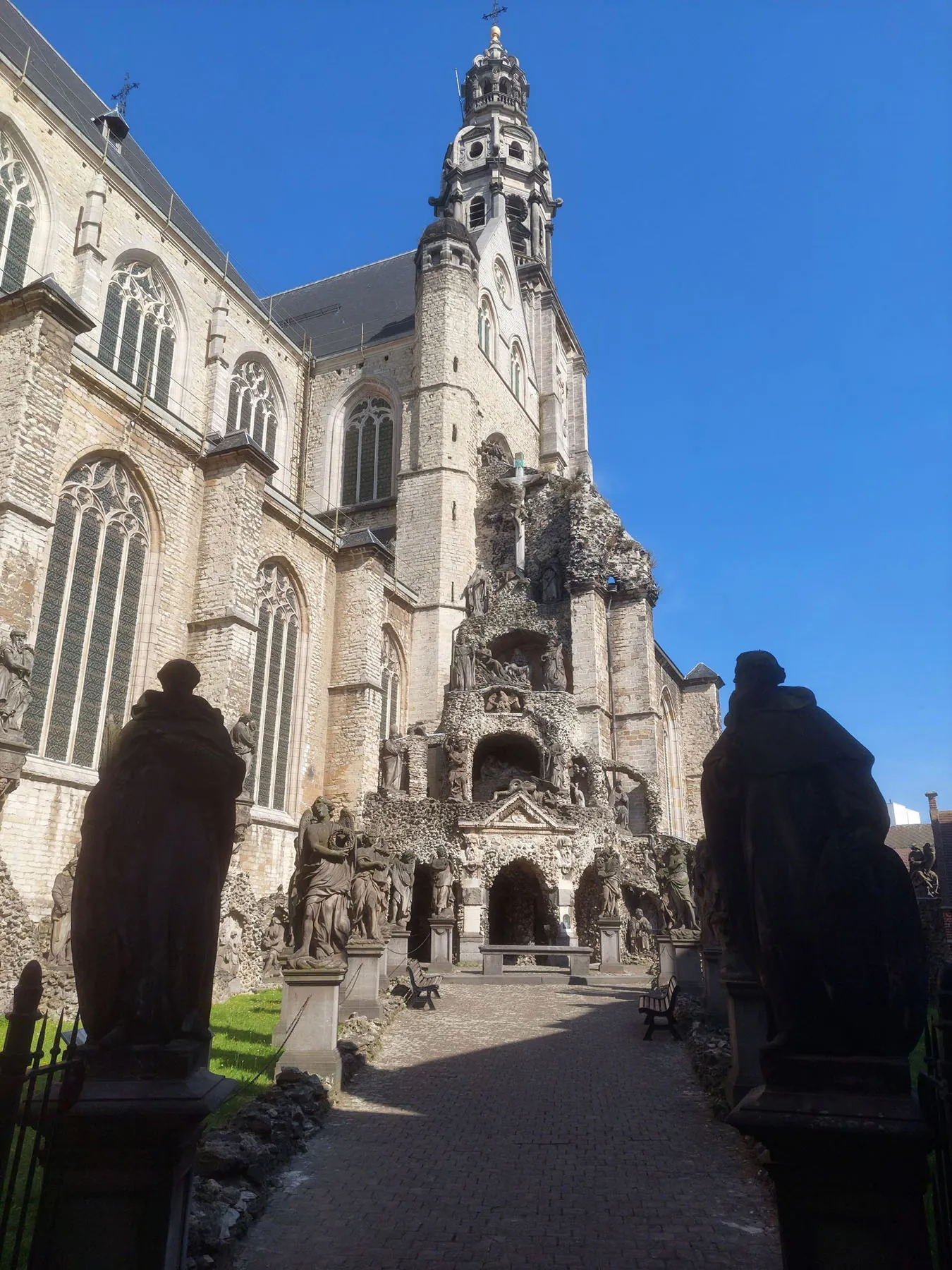 Exterior de una iglesia gótica con una torre barroca alta y ornamentada que se alza sobre un cielo azul despejado, con una gruta de jardín del Calvario y estatuas de piedra en primer plano.