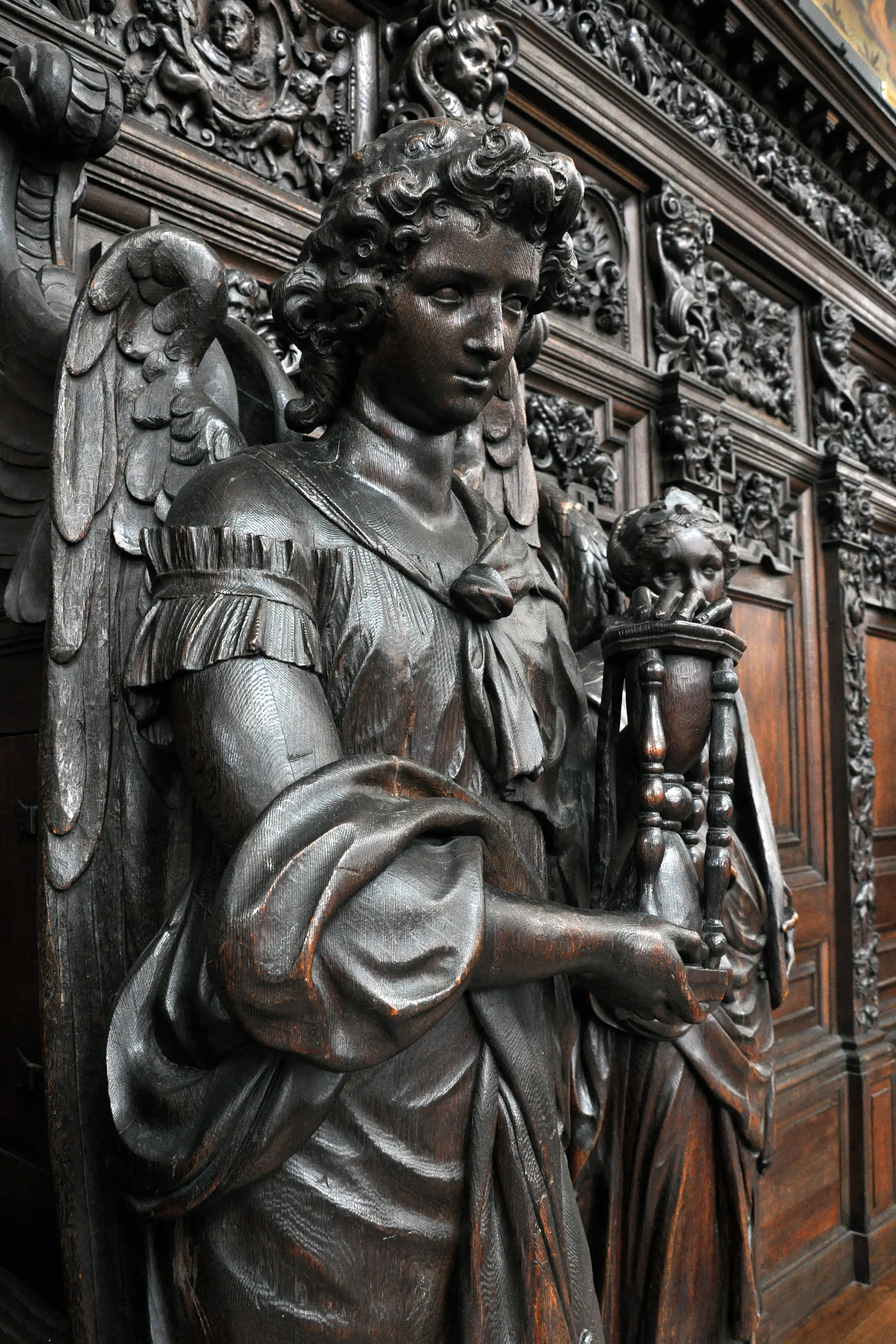 Escultura de madera oscura de un ángel alado sosteniendo una antorcha, sobre paneles de madera tallados con intrincados detalles barrocos.