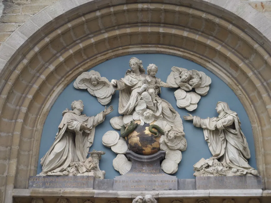 Relieve escultórico blanco de la Virgen María con el niño, junto a santos y ángeles, en un arco semicircular azul sobre una puerta.