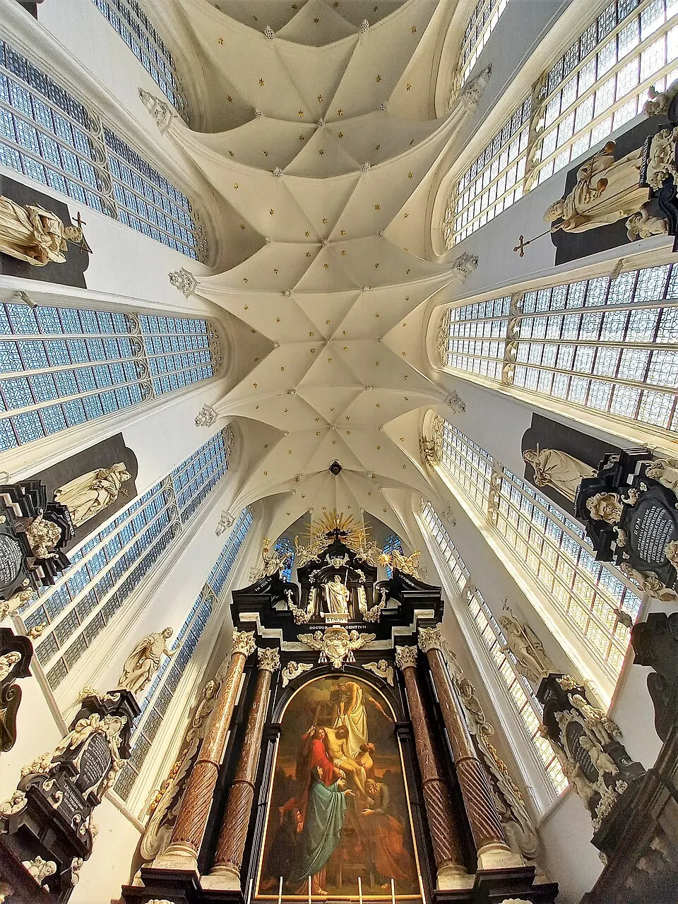 Observando el techo blanco de bóveda de crucería gótica con nervaduras de la Iglesia de San Pablo, con un altar barroco y un gran retablo visibles debajo, flanqueado por altos ventanales arqueados y estatuas en hornacinas.