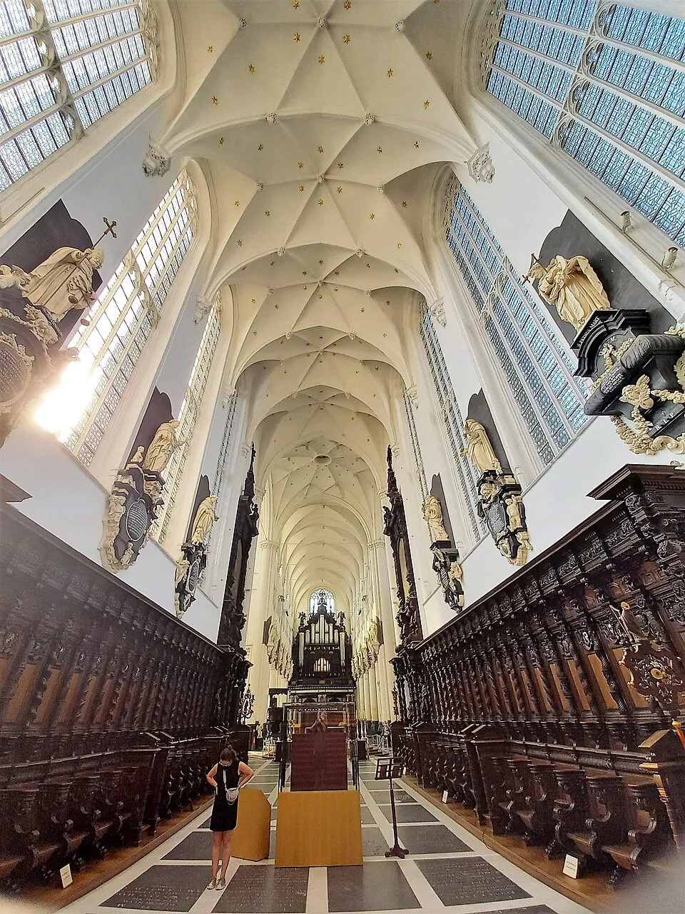 Vista interior de la nave de una iglesia gótica con un techo abovedado blanco, altos vitrales, figuras esculpidas en las paredes y sillería de coro de madera oscura a ambos lados, con un visitante de pie en primer plano.