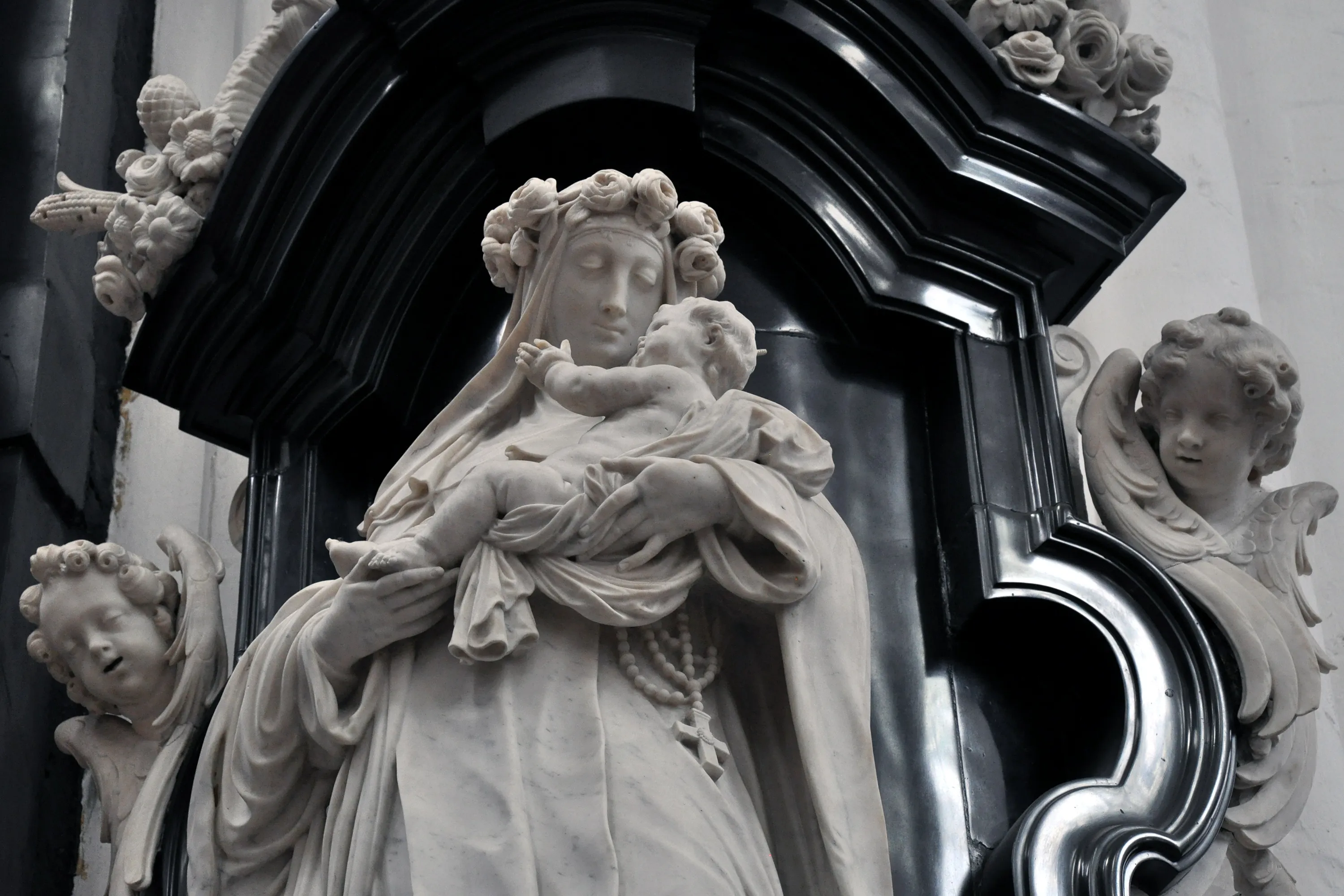 Estatua de mármol blanco de la Virgen María sosteniendo al niño Jesús, flanqueada por figuras de querubines, situada dentro de una oscura y ornamentada hornacina barroca.