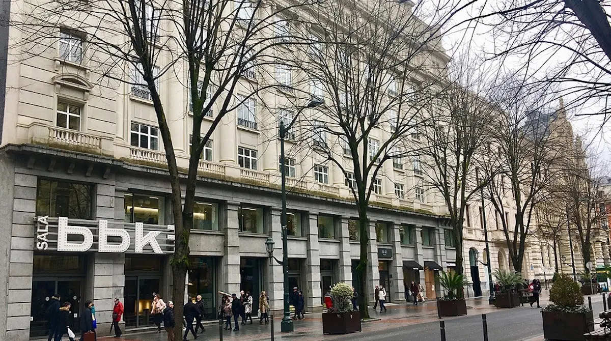 Vista a nivel de calle de la fachada del edificio Sala BBK con cartelería blanca de gran tamaño, árboles sin hojas a lo largo de la acera y peatones caminando frente a la entrada.