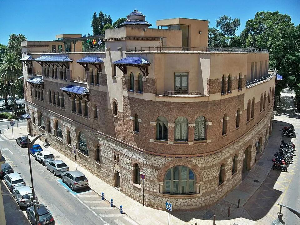 Vista exterior de un gran edificio histórico de ladrillo y piedra con ventanas arqueadas, toldos de azulejos azules y una fachada de esquina curva, con coches aparcados a lo largo de la calle.