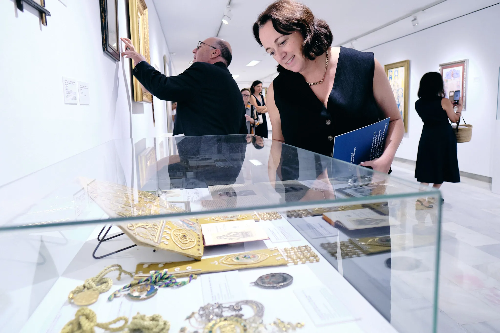 Visitantes observando artefactos en una vitrina de cristal dentro de una galería de exposiciones blanca y luminosa con obras de arte enmarcadas en las paredes.