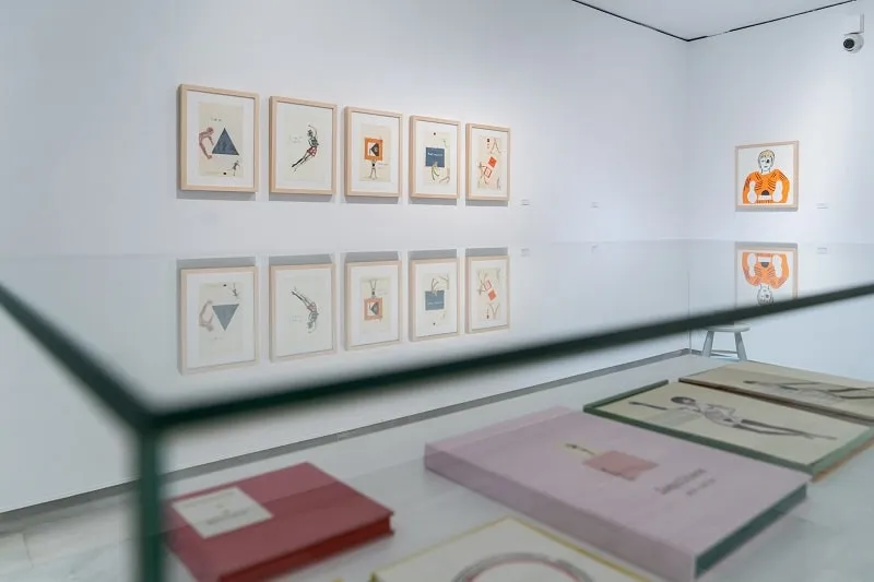 Láminas enmarcadas expuestas en paredes blancas de galería con libros y documentos en una vitrina de cristal en primer plano