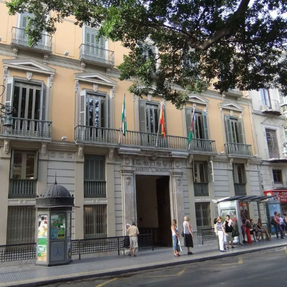Vista exterior de una fachada de edificio clásico de color amarillo con balcones, barandillas de hierro, banderas sobre la entrada y personas en la acera.