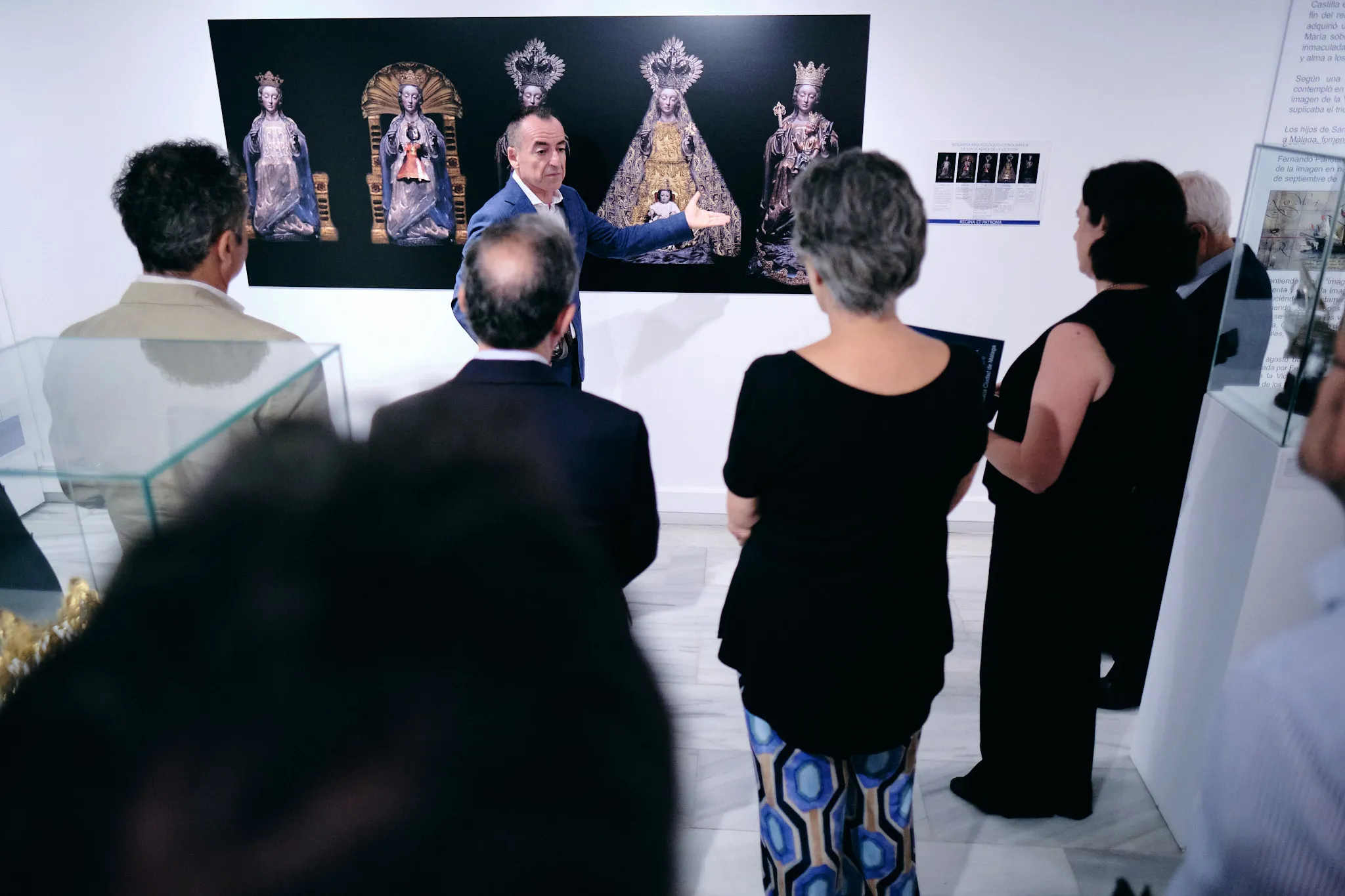 Grupo de visitantes observando un gran panel de exposición con cuatro figuras religiosas coronadas en una galería, mientras un guía hace una presentación al público.