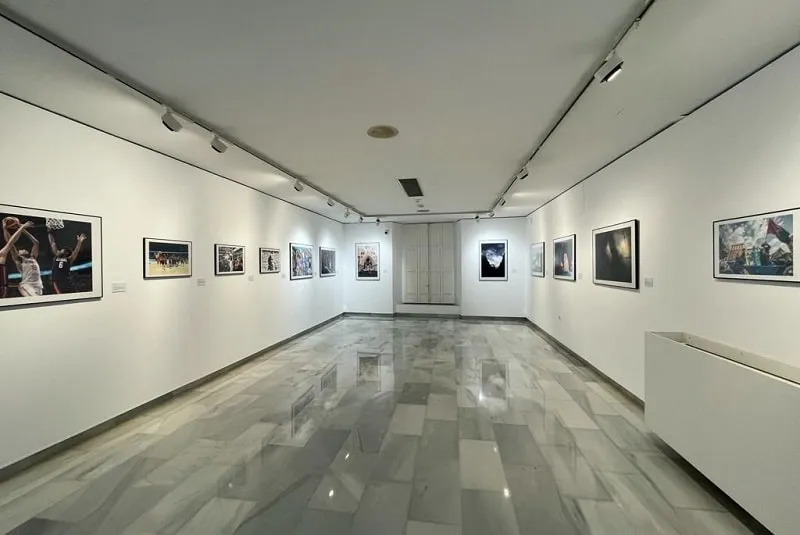Una larga sala de exposiciones blanca con fotografías enmarcadas colgadas en ambas paredes y un suelo de baldosas brillantes que refleja las luces del techo.