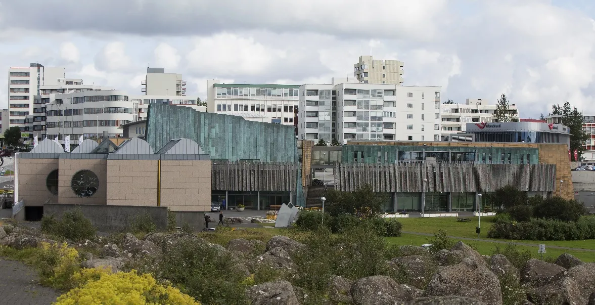 Vista exterior de la sala de conciertos Salurinn que presenta una fachada curva de piedra con una gran ventana circular, una sección alta de cristal verde y un ala de listones de madera, con edificios de apartamentos blancos y un terreno rocoso en primer plano.