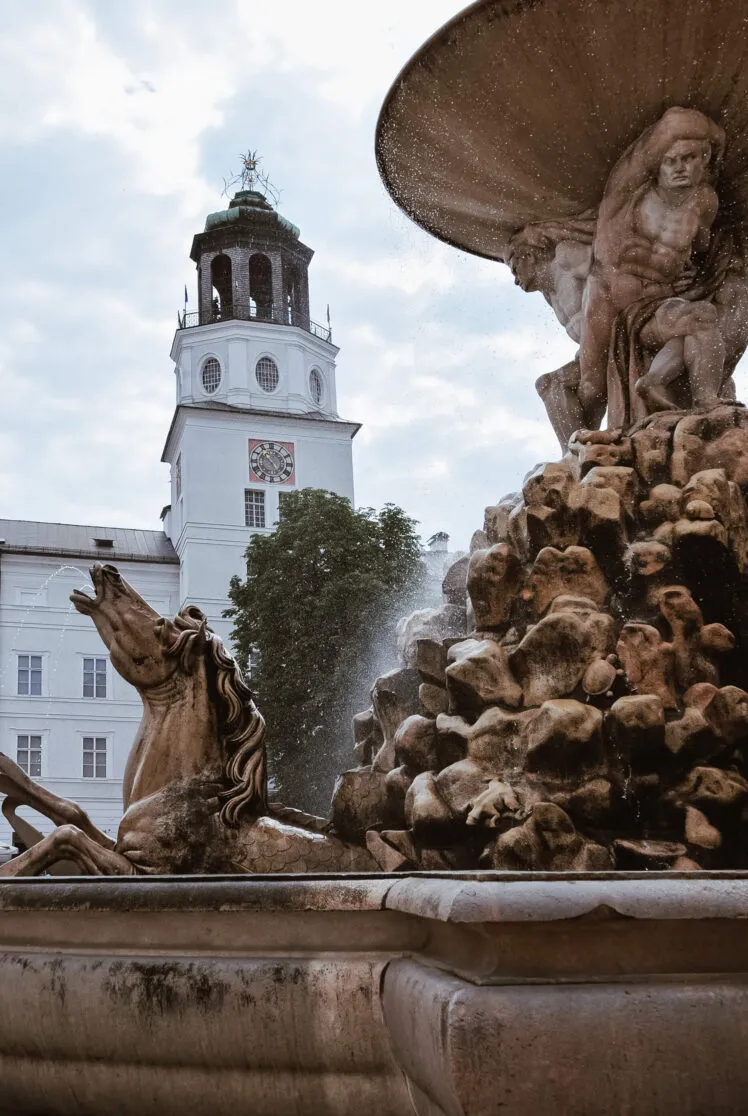 Stone fountain sculptures with a rearing horse and mythological figures in the foreground, and the white Neue Residenz tower with its green-domed carillon belfry rising behind it.
