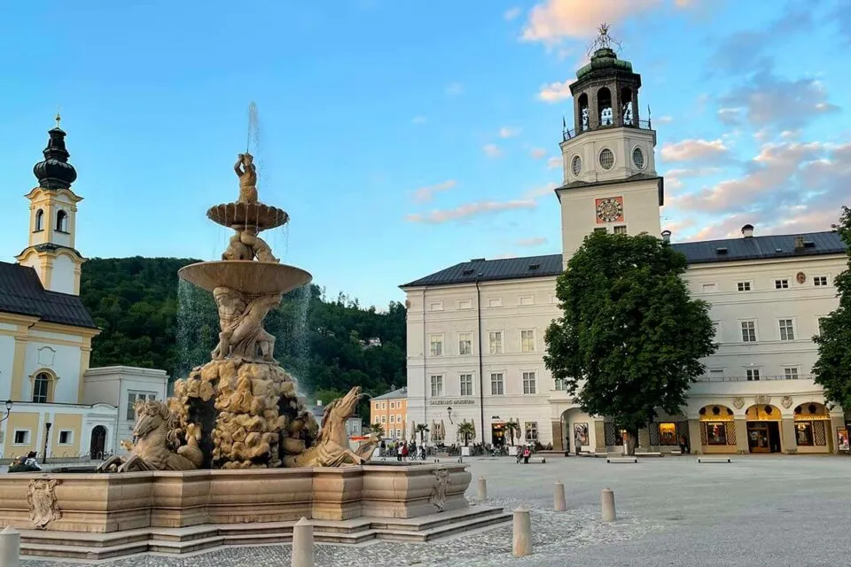 Wide view of Salzburg's Residenzplatz with the elaborate multi-tiered Residence Fountain in the foreground and the white Neue Residenz building with its clock tower on the right under a blue evening sky.