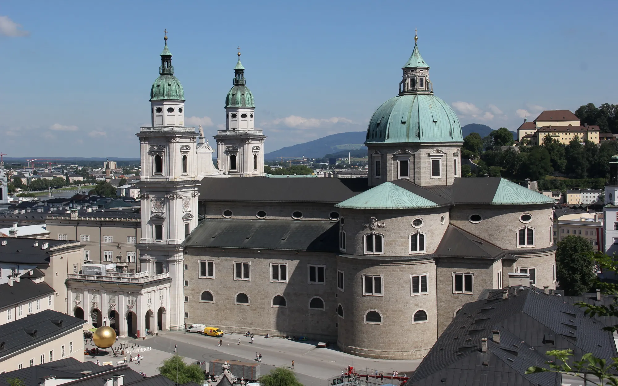 Widok z lotu ptaka na Katedrę w Salzburgu, ukazujący bliźniacze wieże z miedzianymi iglicami w kolorze zieleni, dużą centralną kopułę oraz otaczającą panoramę miasta z górami w tle.