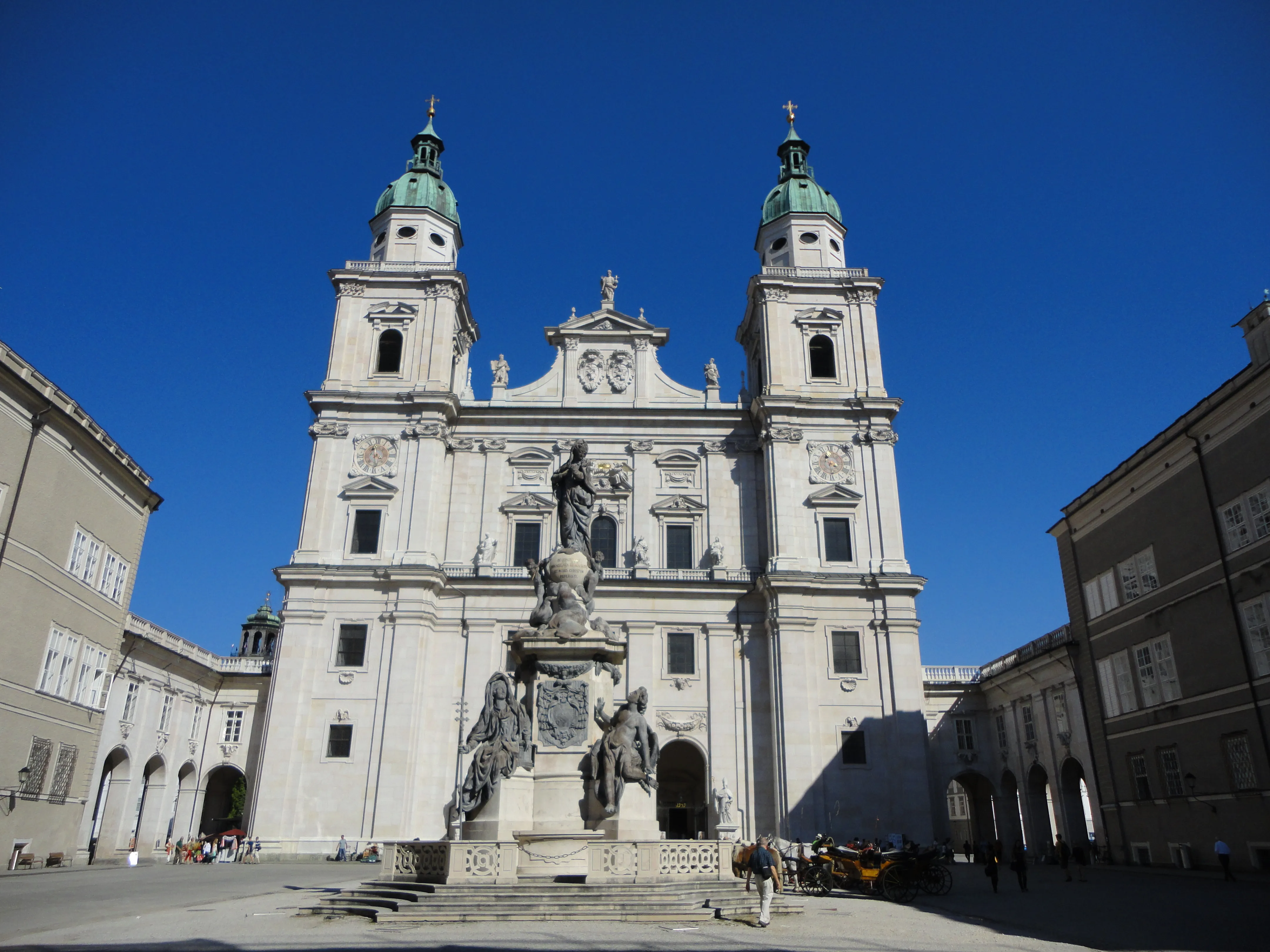 Biała barokowa fasada katedry w Salzburgu z dwiema wieżami z zielonymi kopułami i centralną kolumną z posągiem na pierwszym planie na tle bezchmurnego błękitnego nieba