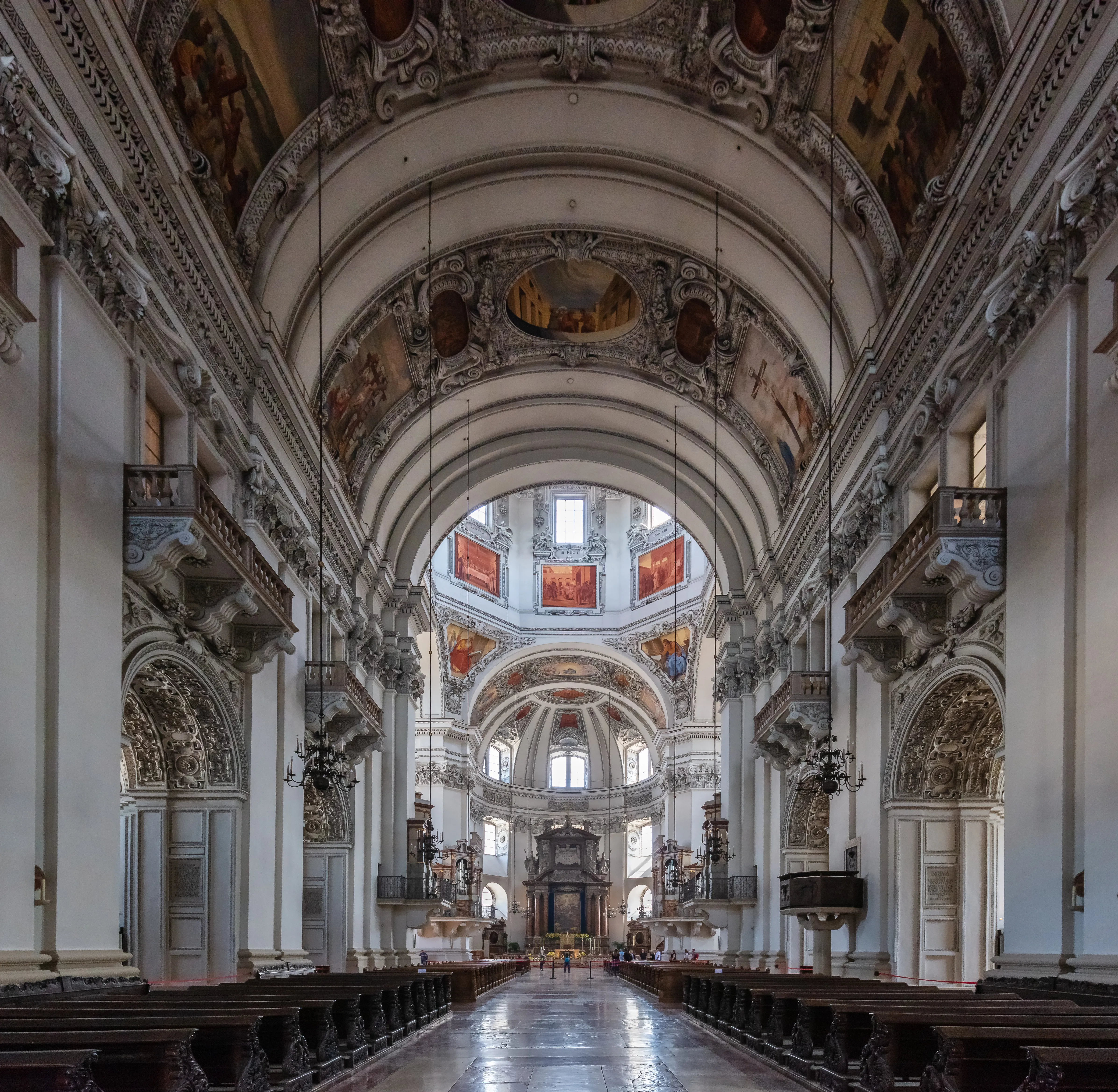 Wnętrze katedry w Salzburgu ukazujące sklepienie nawy głównej z barokowymi freskami, zdobione kolumny, drewniane ławki oraz widoczną w oddali kopułę.