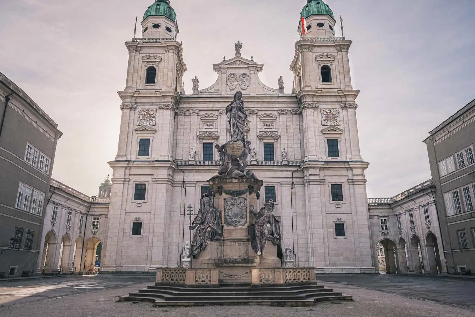 Barokowa fasada katedry w Salzburgu z bliźniaczymi wieżami zwieńczonymi zielonymi miedzianymi kopułami oraz fontanną Marienbrunnen na placu na pierwszym planie.