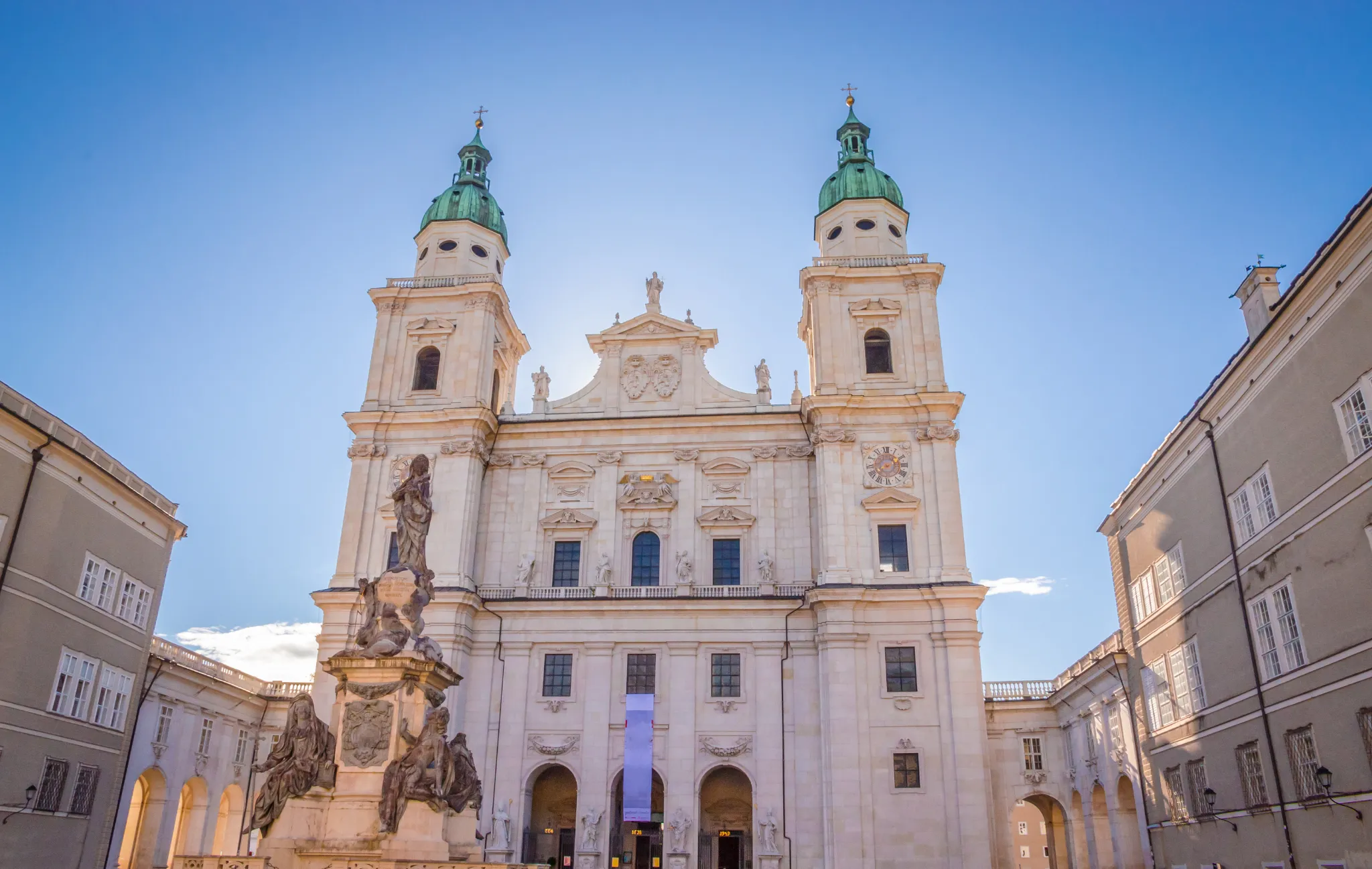 Fasada katedry w Salzburgu z dwiema wieżami z miedzianymi kopertami, barokową architekturą oraz kamienną fontanną na pierwszym planie na tle błękitnego nieba