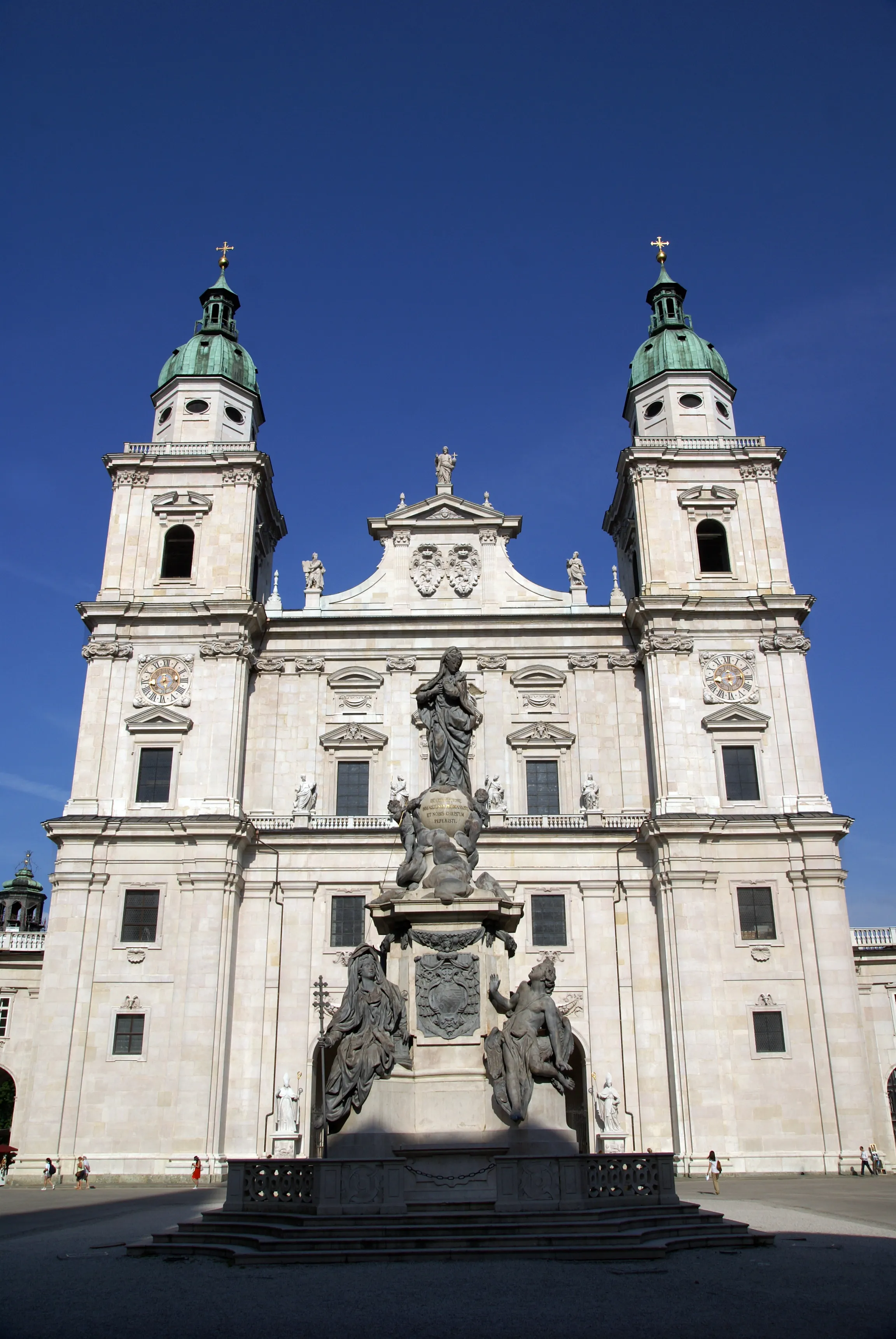 Biała barokowa fasada katedry w Salzburgu z dwiema wieżami z zielonymi kopułami flankującymi centralny szczyt, z rzeźbioną fontanną na pierwszym planie na tle bezchmurnego błękitnego nieba