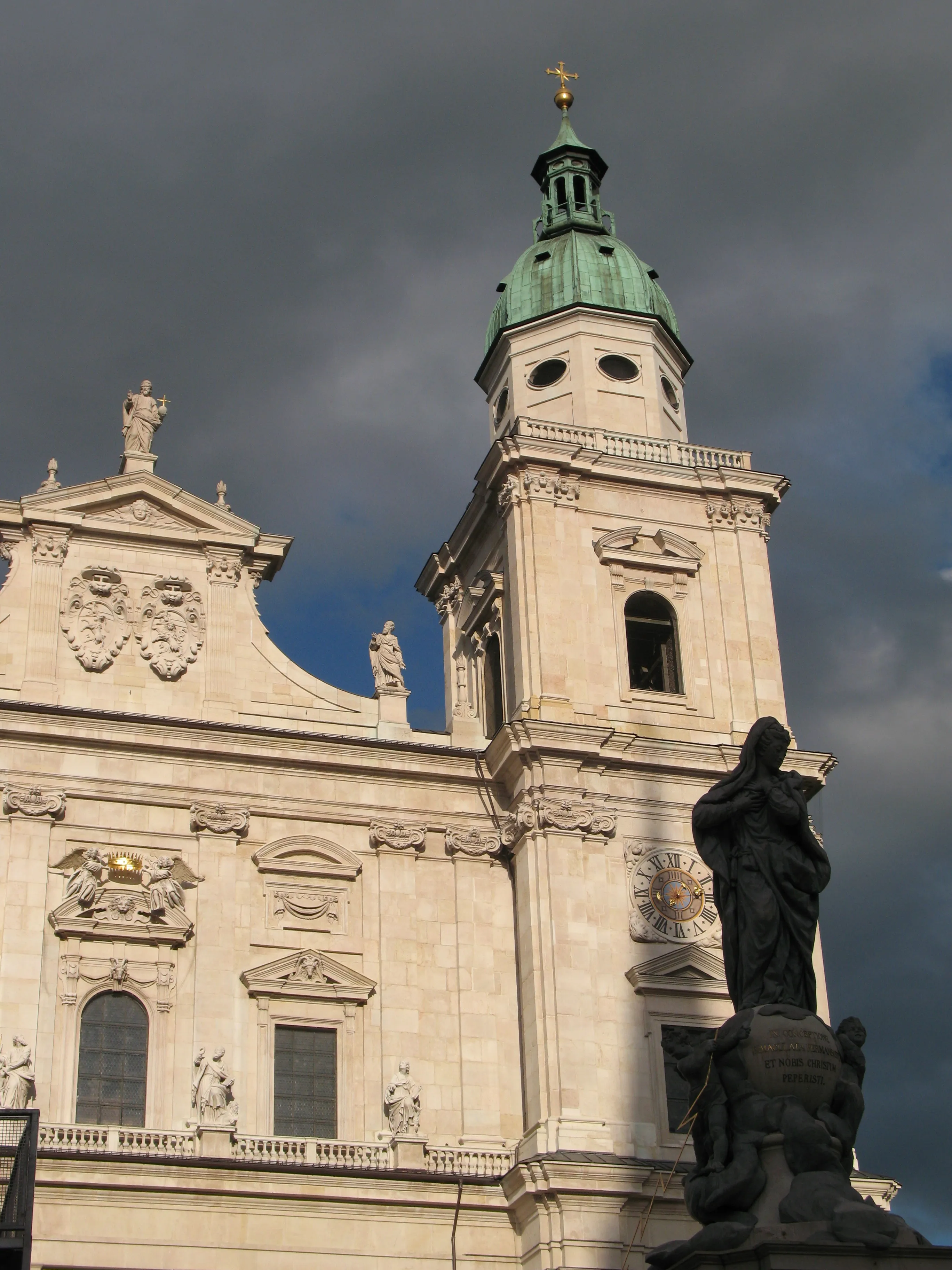Fachada barroca de la Catedral de Salzburgo con una torre de cúpula de cobre verde rematada por una cruz dorada, y una estatua de bronce oscuro en primer plano contra un cielo tormentoso