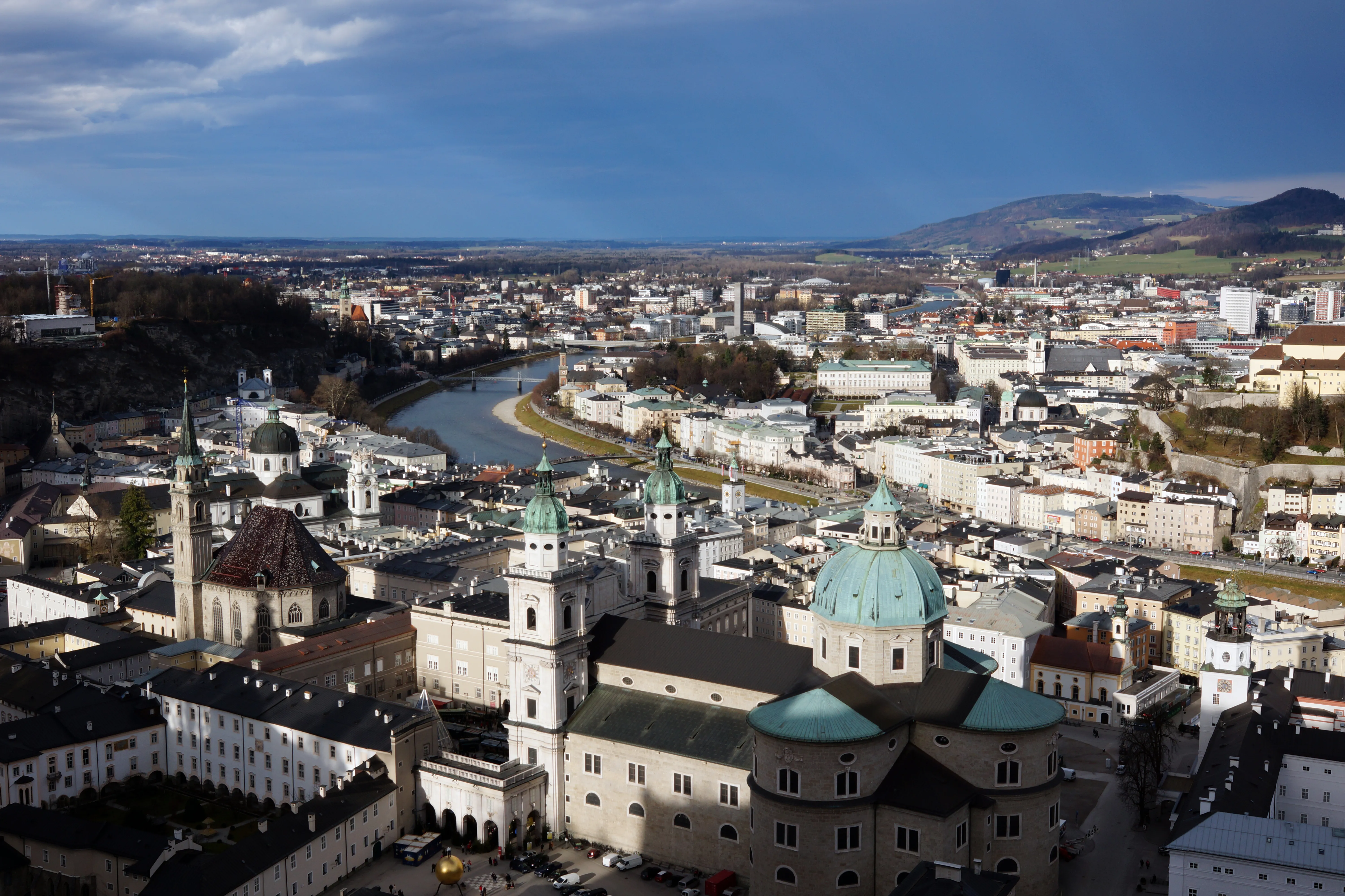 Szerokie ujęcie z lotu ptaka na zielone kopuły i wieże katedry w Salzburgu, z rzeką Salzach wijącą się przez miasto i wzgórzami w tle.