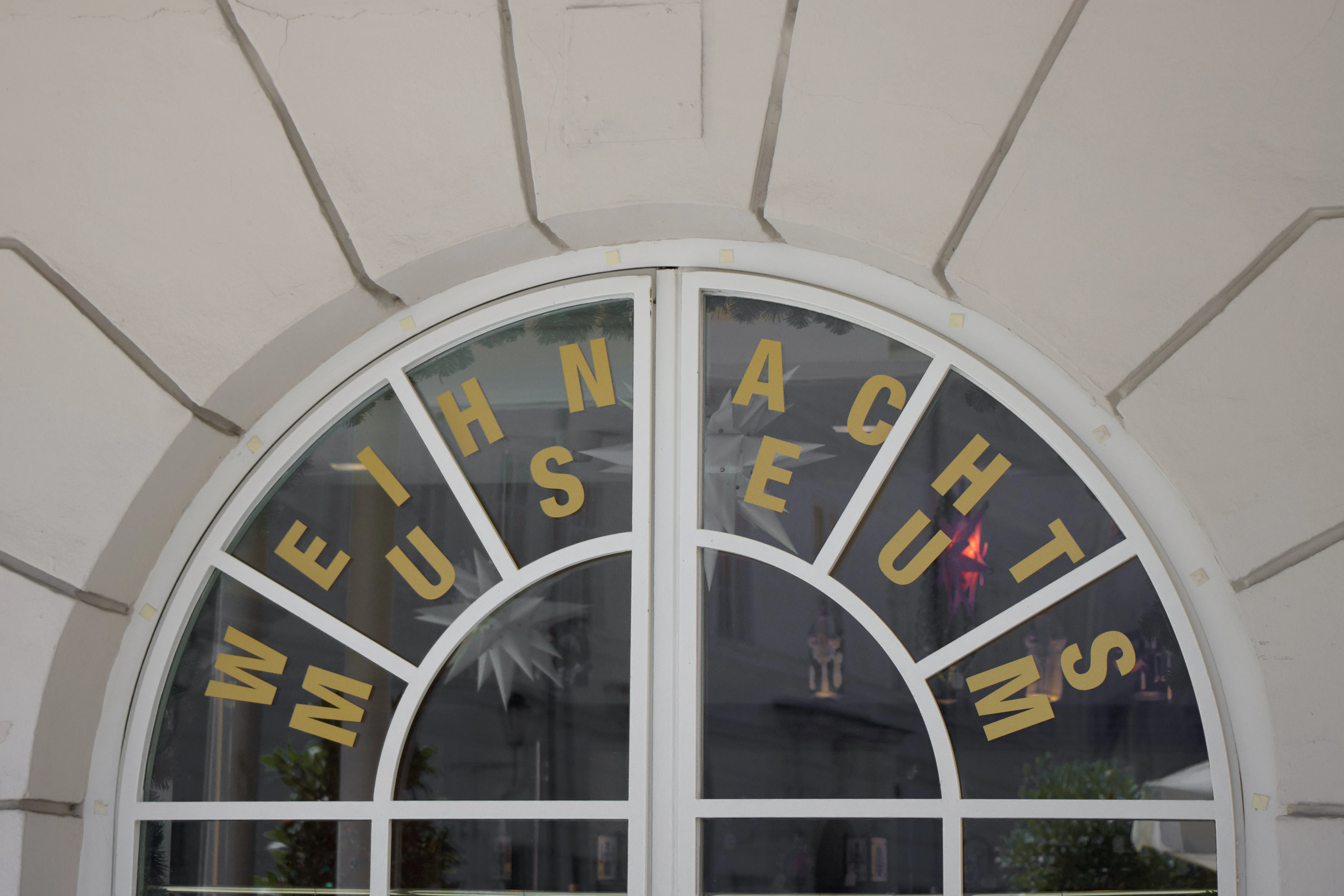 Ventana arqueada con marco blanco y letras doradas que dicen "WEIHNACHTS MUSEUM" dispuestas en forma de abanico sobre los cristales oscuros.