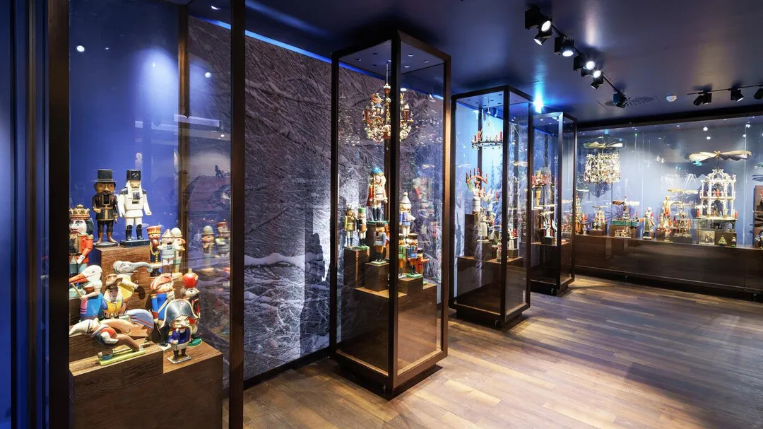 Interior de museo con una fila de vitrinas de cristal llenas de cascanueces y objetos festivos bajo iluminación azul de acento y luces de riel.