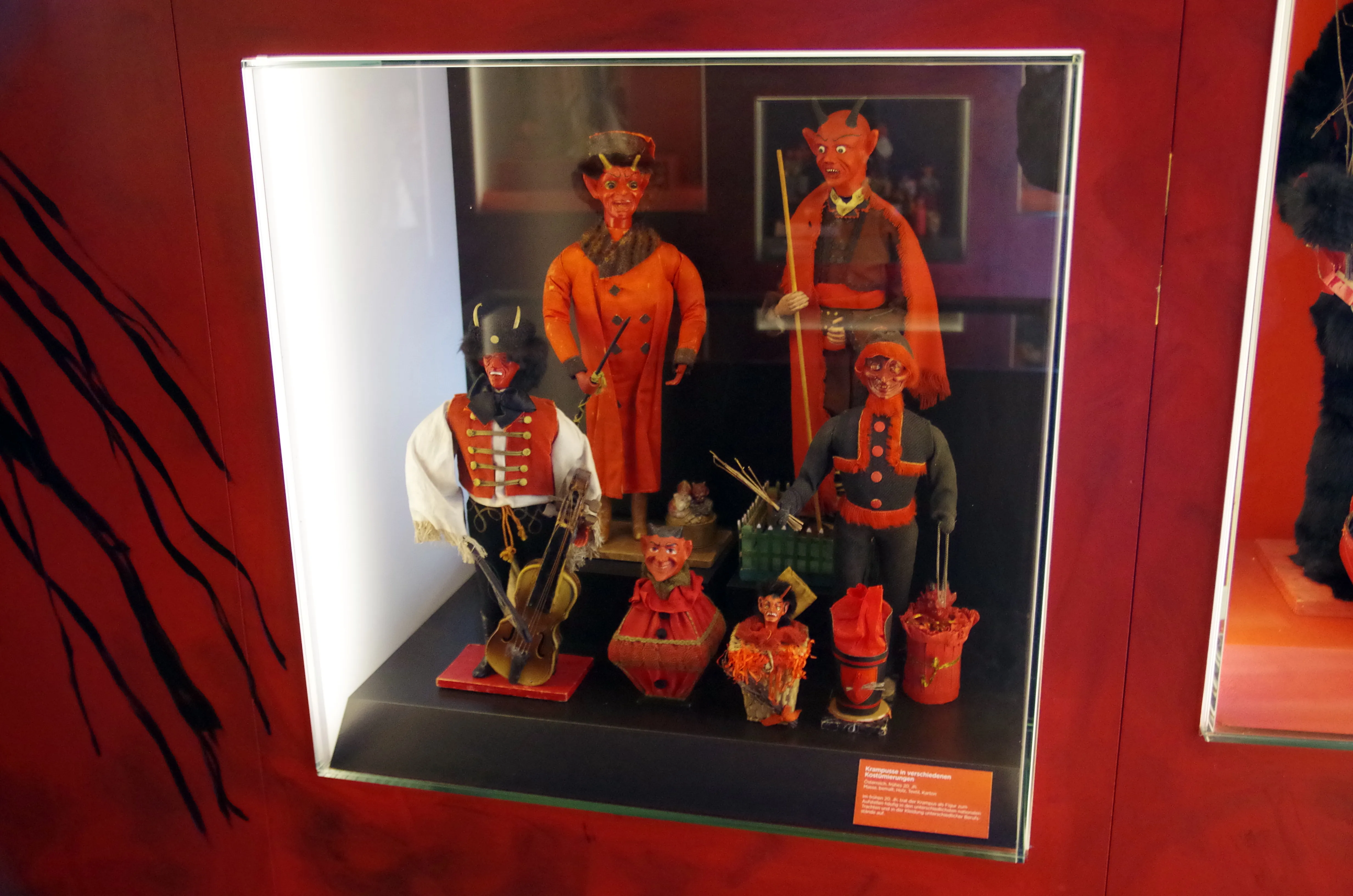 Vitrina de museo que contiene figuras tradicionales de Krampus y Perchten con trajes coloridos colocadas sobre una pared roja