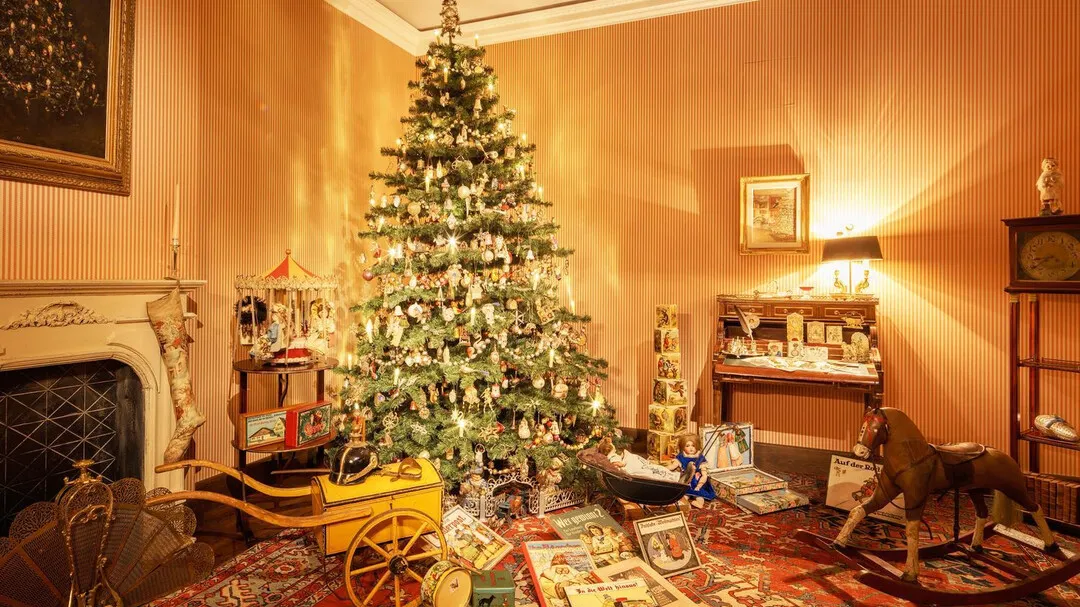 Una sala de museo con iluminación cálida, adornada con un árbol de Navidad, una chimenea blanca, un carruaje de juguete amarillo, un caballito balancín de madera y adornos navideños vintage dispuestos sobre una alfombra estampada.