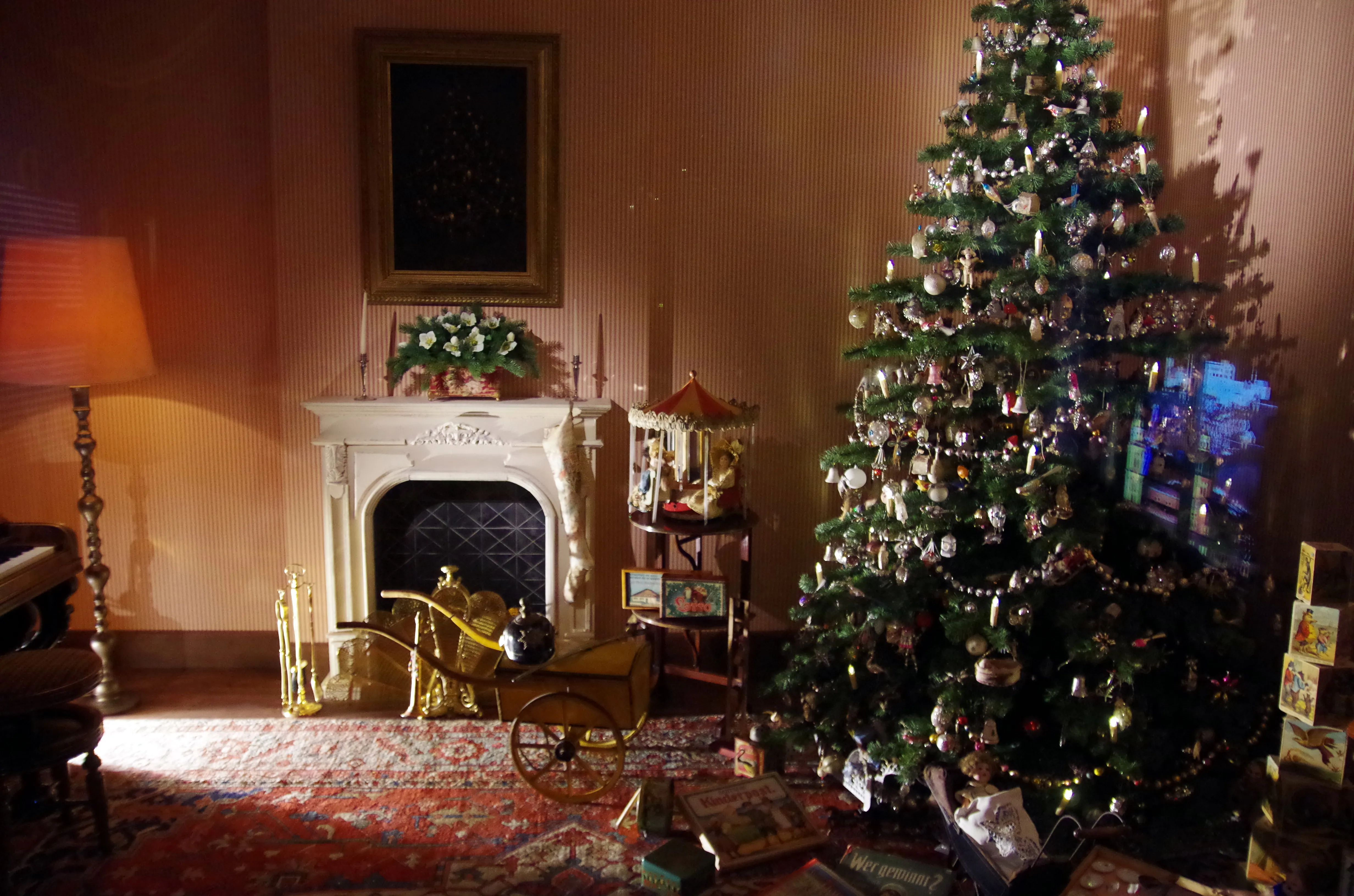 Sala de exposición de museo con un árbol de Navidad decorado, una chimenea blanca con arreglo floral, juguetes antiguos y luz cálida de lámpara sobre una alfombra estampada