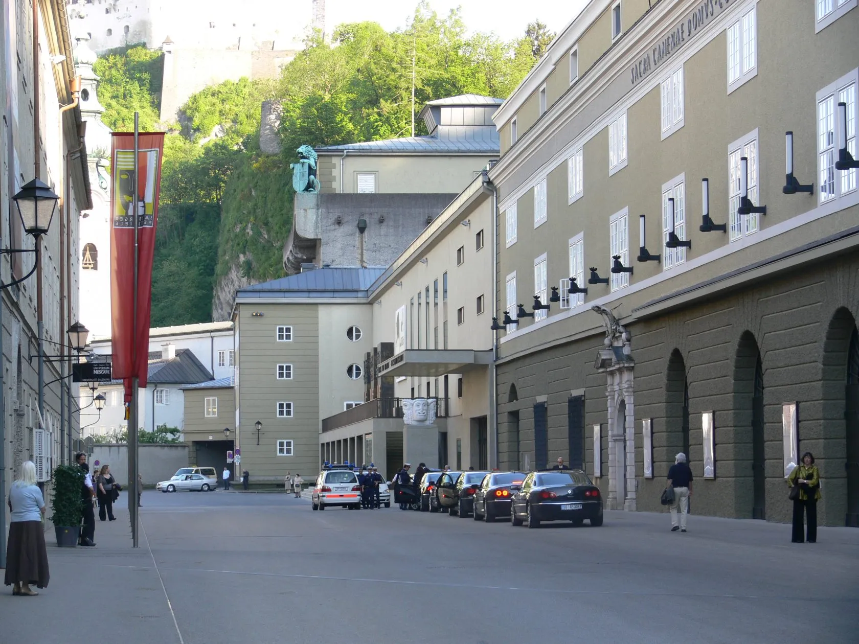 Straatzicht op de Salzburg Festival Halls met het gebouw met de booggevel aan de rechterkant, geparkeerde auto's, voetgangers en Hohensalzburg Fortress zichtbaar op de heuvel op de achtergrond