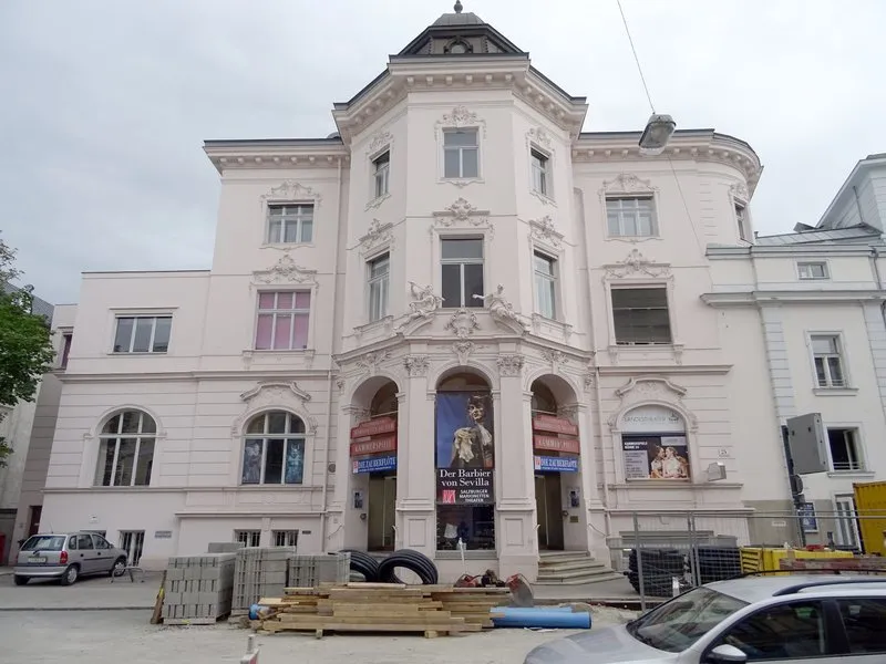 Fachada blanca del edificio histórico del Salzburg Marionette Theatre con pórtico de entrada arqueado, ornamentación en piedra, torre de esquina con cúpula y materiales de construcción apilados frente a la entrada