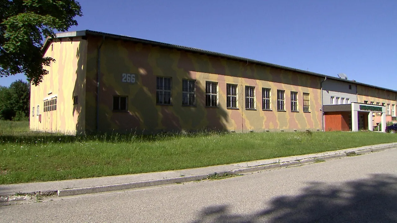 Long single-story barrack building painted in yellow and brown camouflage pattern with the number 266 on its wall, multiple windows, green lawn, and paved road in front under a blue sky