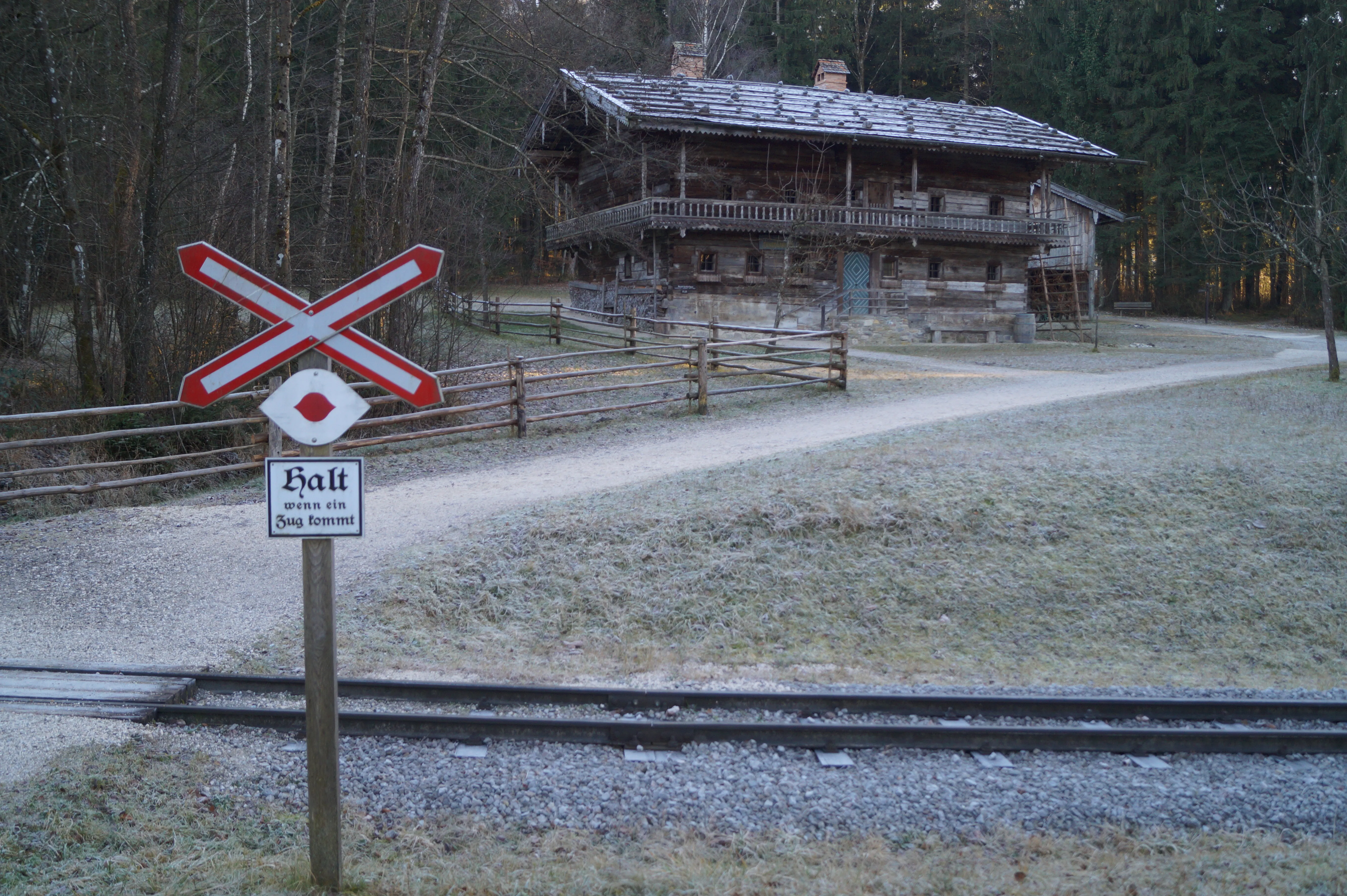Een rood-wit spoorwegovergangsbord met het opschrift "Halt" staat naast smalspoorrails met een traditionele houten boerderij in de Alpen op de achtergrond.