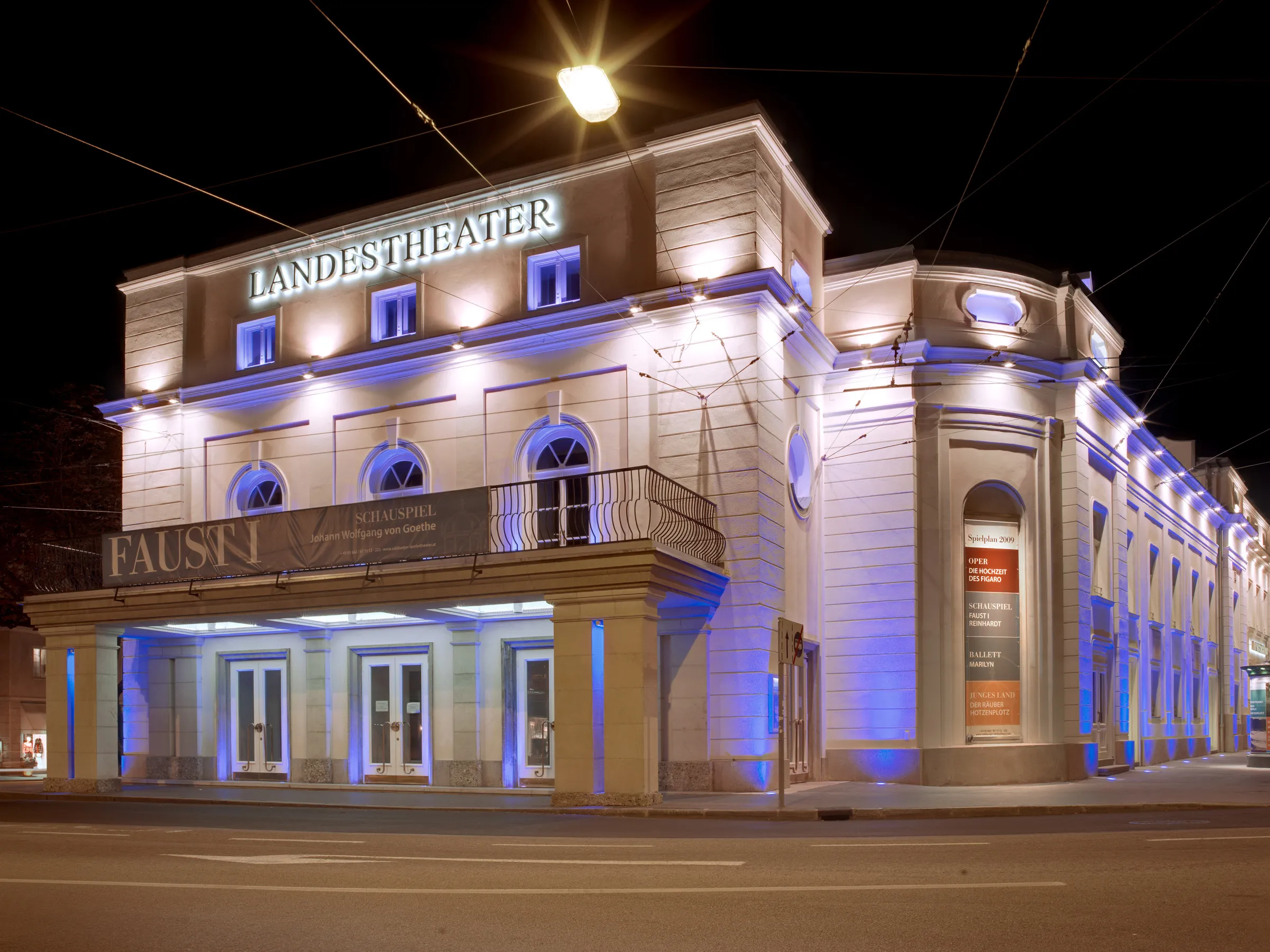 Nighttime exterior view of the Salzburger Landestheater building illuminated with blue and white lights, featuring large 'LANDESTHEATER' signage on the facade.